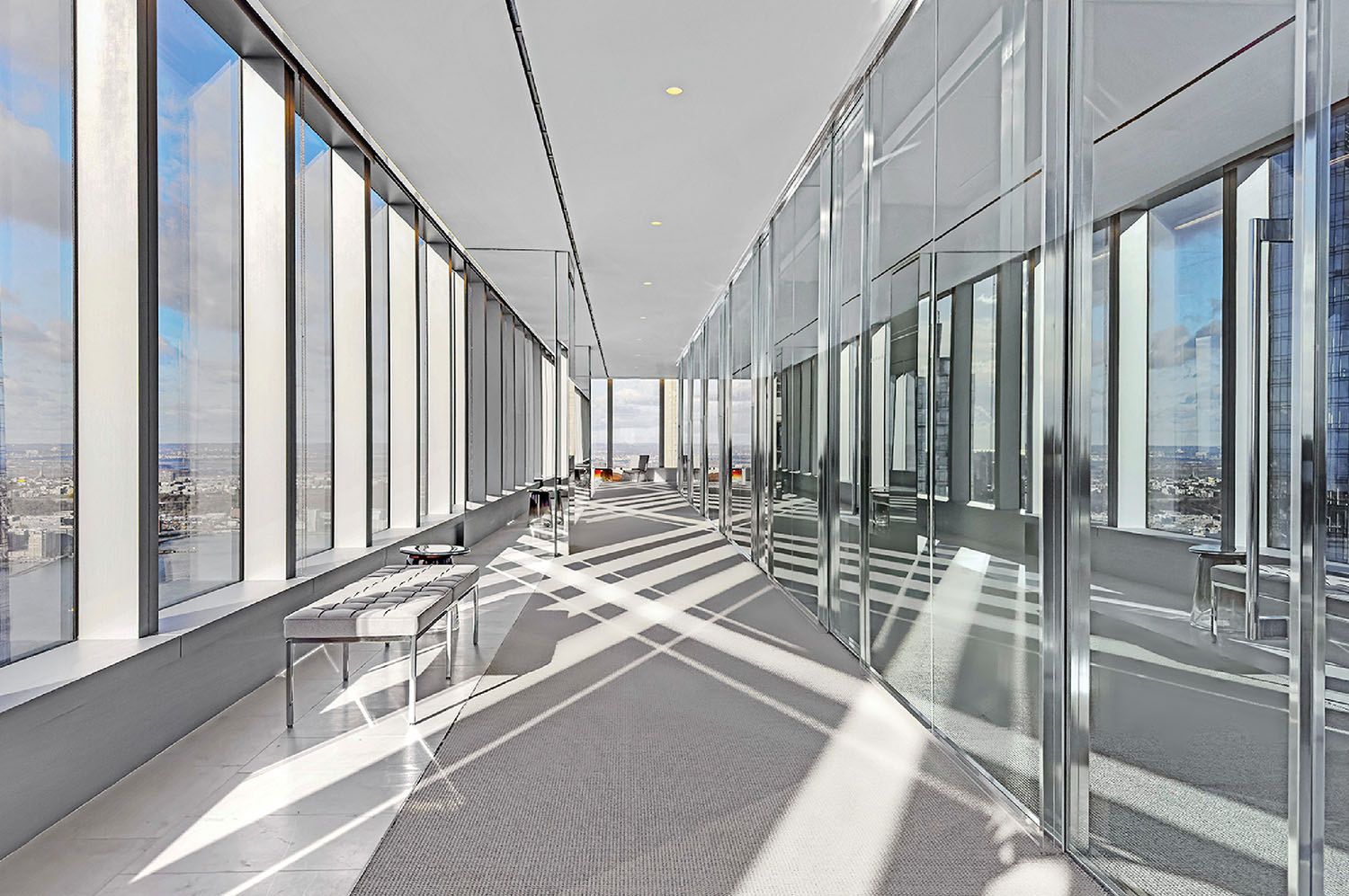 A modern hallway with floor-to-ceiling windows, benches, and glass walls reflecting sunlight on the floor, overlooking a cityscape.