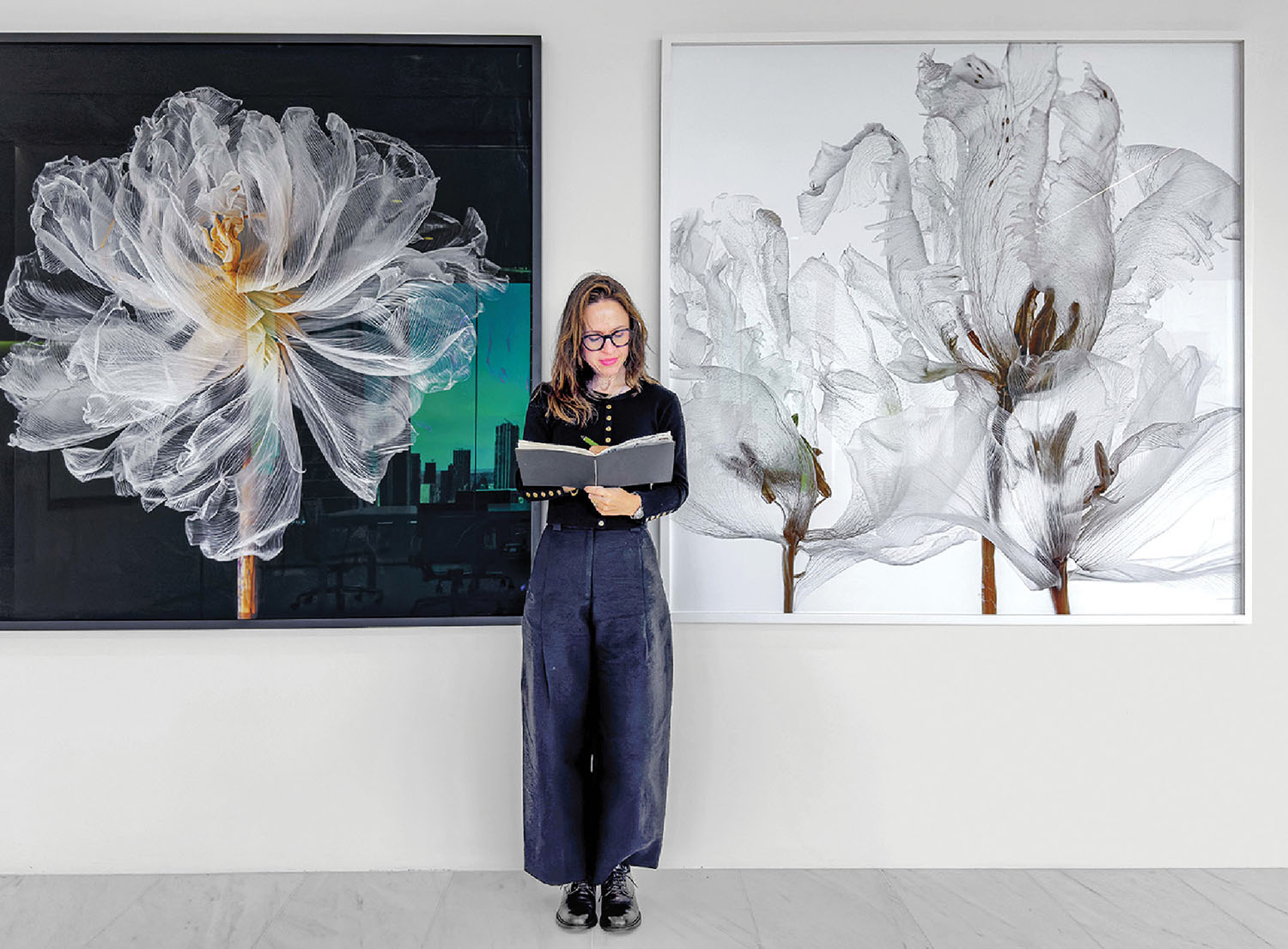 A woman stands reading a notebook between two large framed floral artworks displayed on a white wall.