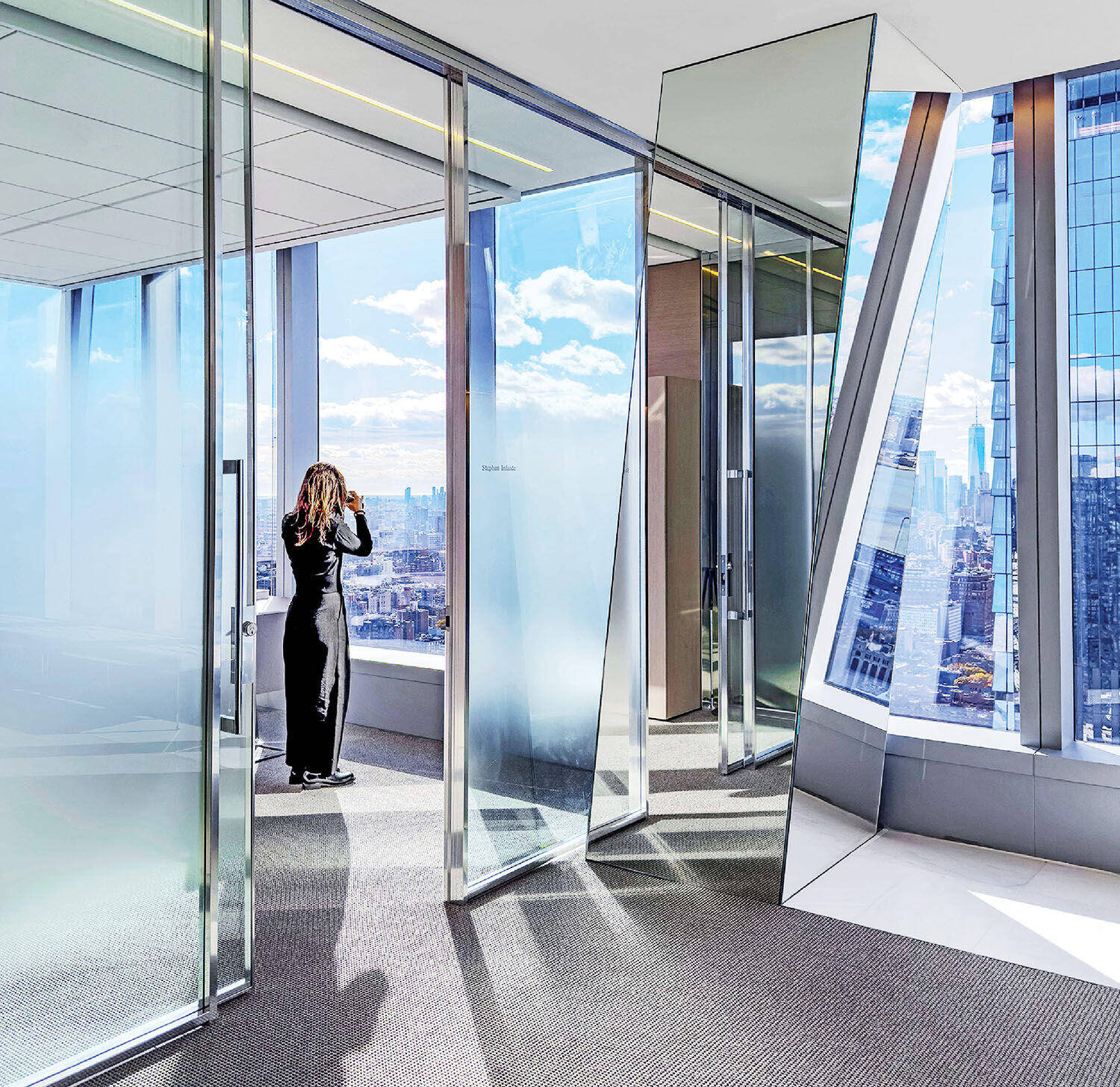 A woman stands by floor-to-ceiling windows in a modern office, looking out at a city skyline on a bright day.