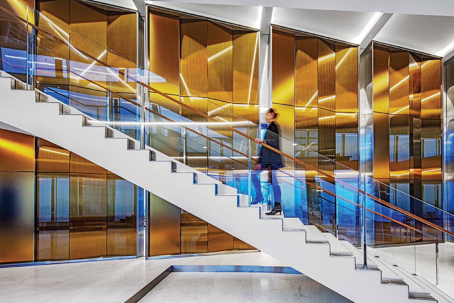A person in business attire walks up a modern white staircase with glass railings, in front of gold-tinted glass wall panels.