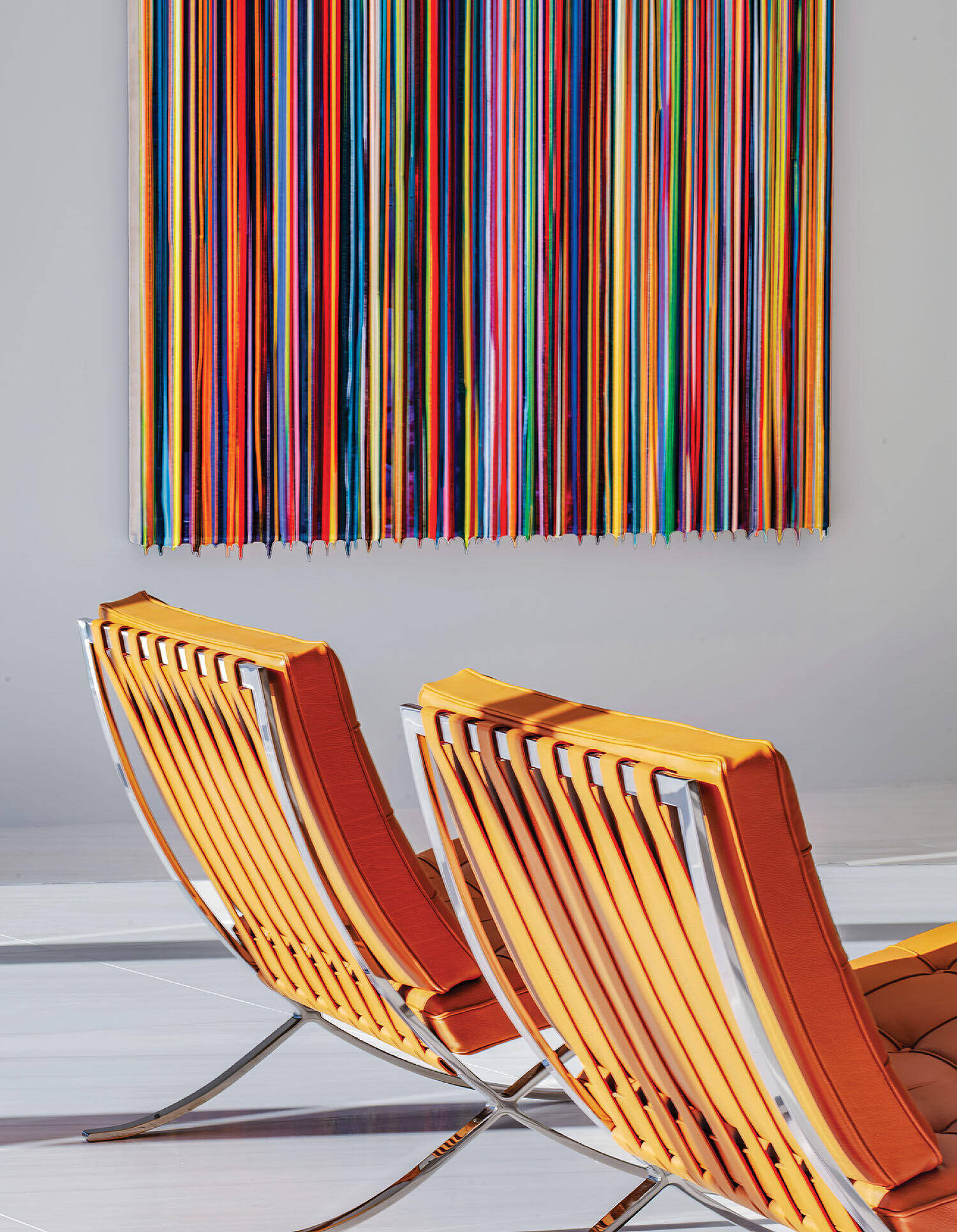 Two orange modern chairs with metal frames are placed in front of a wall displaying a colorful, vertical striped abstract artwork.