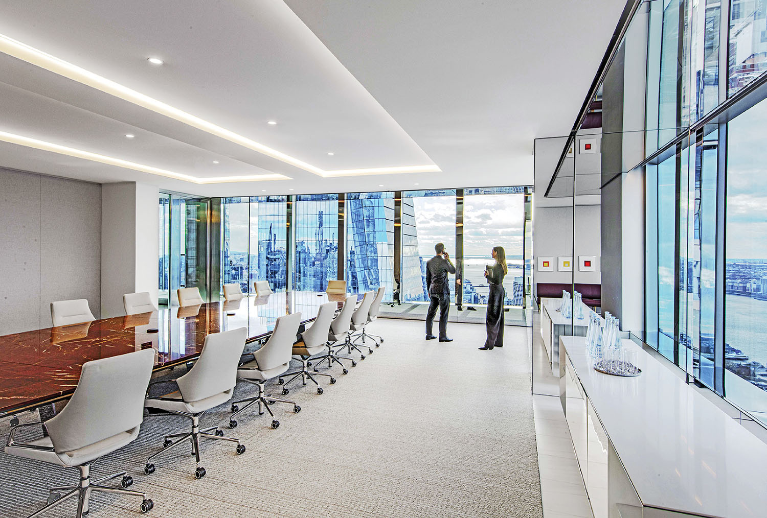 Modern conference room with a long table and white chairs; two people stand near floor-to-ceiling windows overlooking a cityscape.