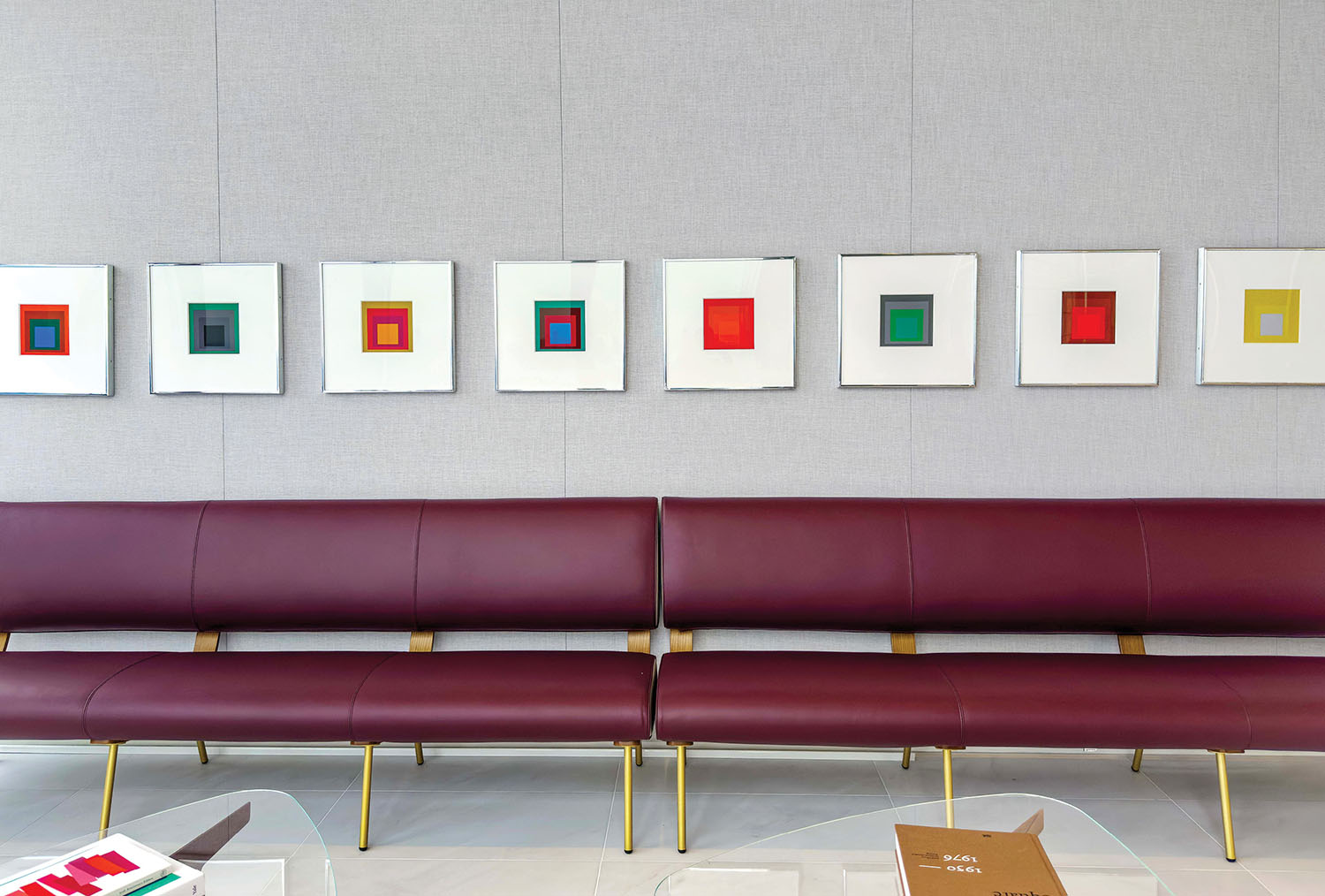 A modern waiting area with maroon benches, glass tables, and a row of framed abstract square artworks on a light gray wall.
