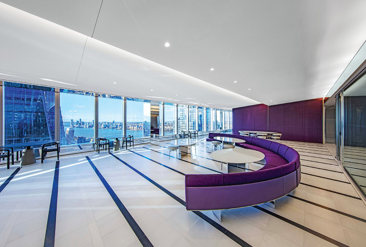 Modern office lounge with purple curved seating, round white tables, and floor-to-ceiling windows overlooking a city skyline and river.