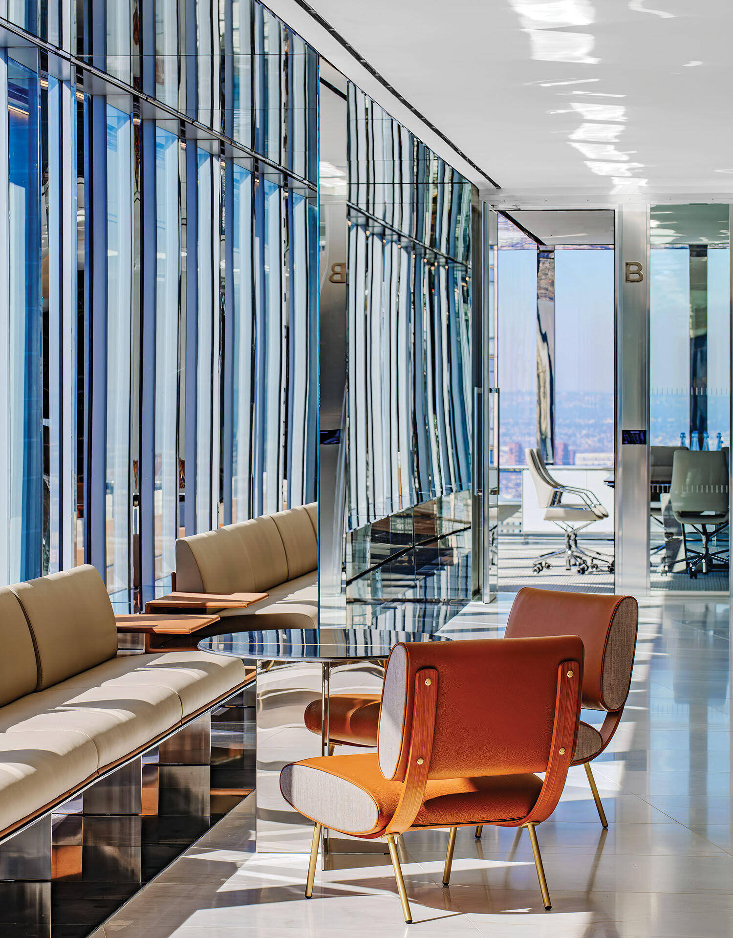 Modern office lounge with beige sofas, orange armchairs, and a glass wall reflecting sunlight, leading to a glass-walled conference room in the background.