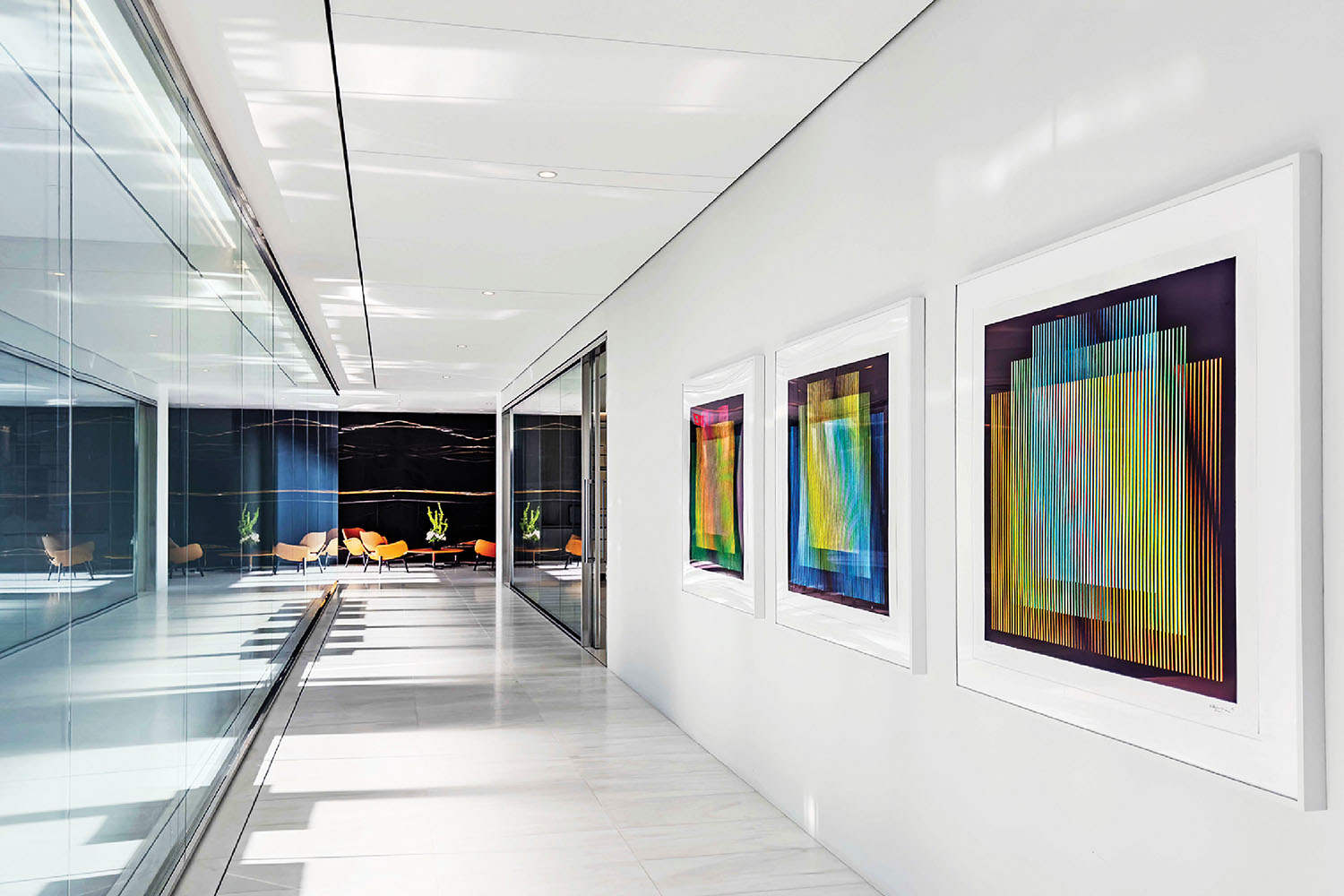 A modern hallway with white floors and walls, featuring three colorful abstract artworks on one wall and glass panels on the opposite side.