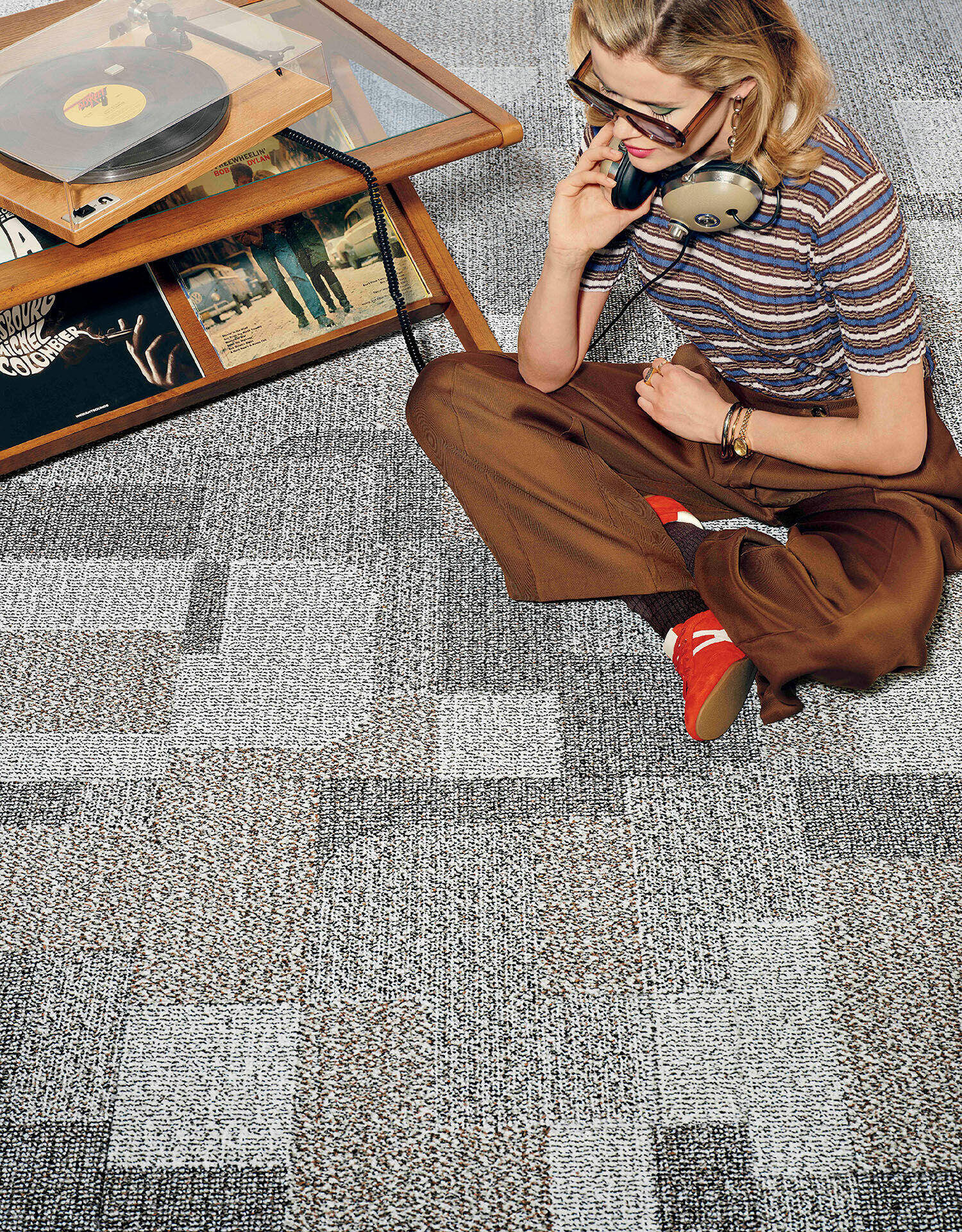 A person sits on a patterned carpet next to a vinyl record player and records, wearing headphones and brown pants with red socks.