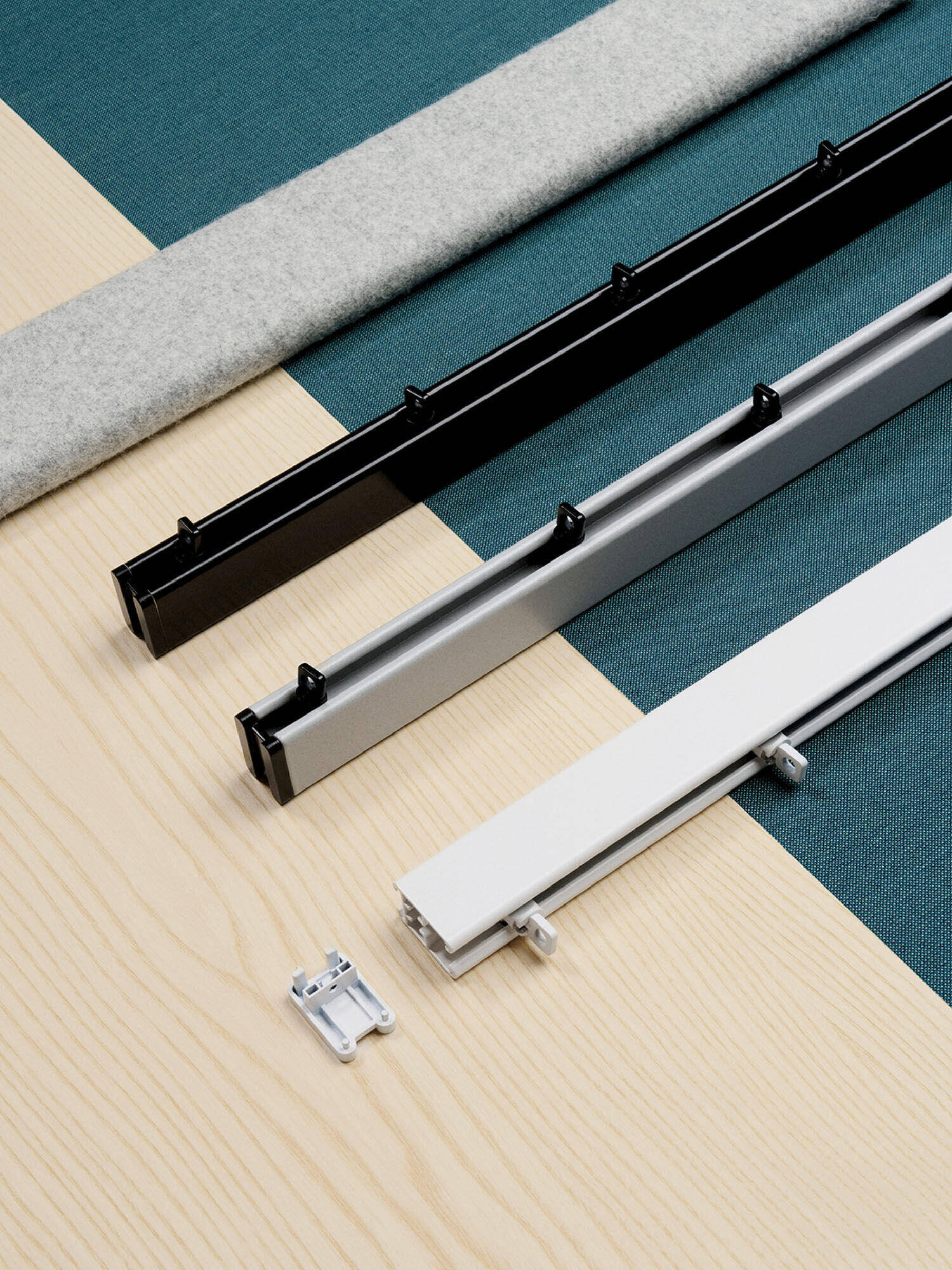 Three horizontal metal rails—black, silver, and white—are arranged parallel on a light wood surface with a gray felt strip and a small metal bracket nearby.
