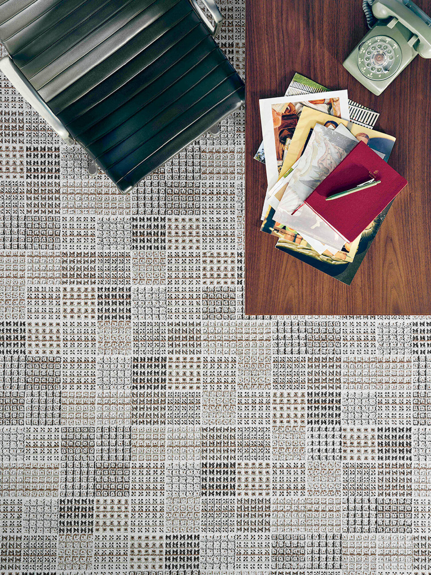 Black office chair and wooden desk with a stack of papers, a red notebook, a pen, and a rotary phone on a patterned tile floor.