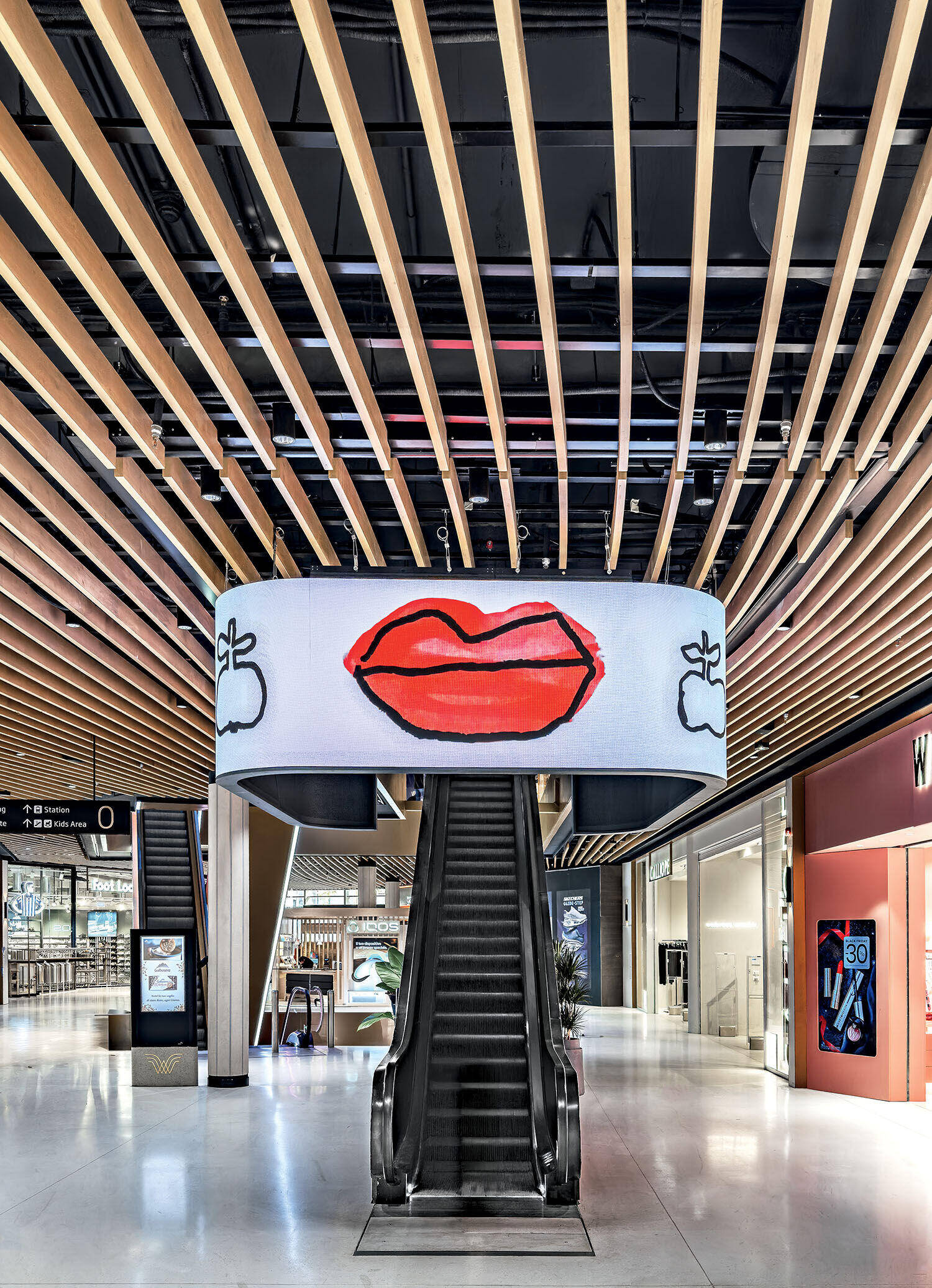 An empty escalator in a modern shopping mall, with a large digital screen above displaying an illustration of red lips and two small apples on each side.