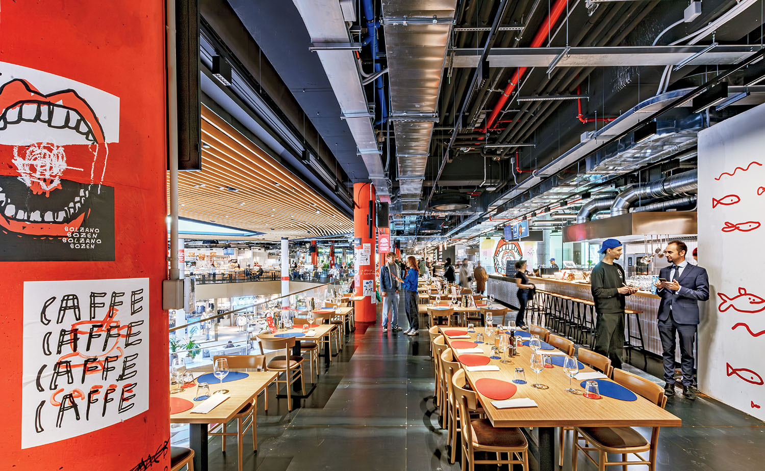 Modern indoor dining area with wooden tables, red chairs, bar seating, and people conversing; colorful wall art and exposed ceiling pipes are visible.