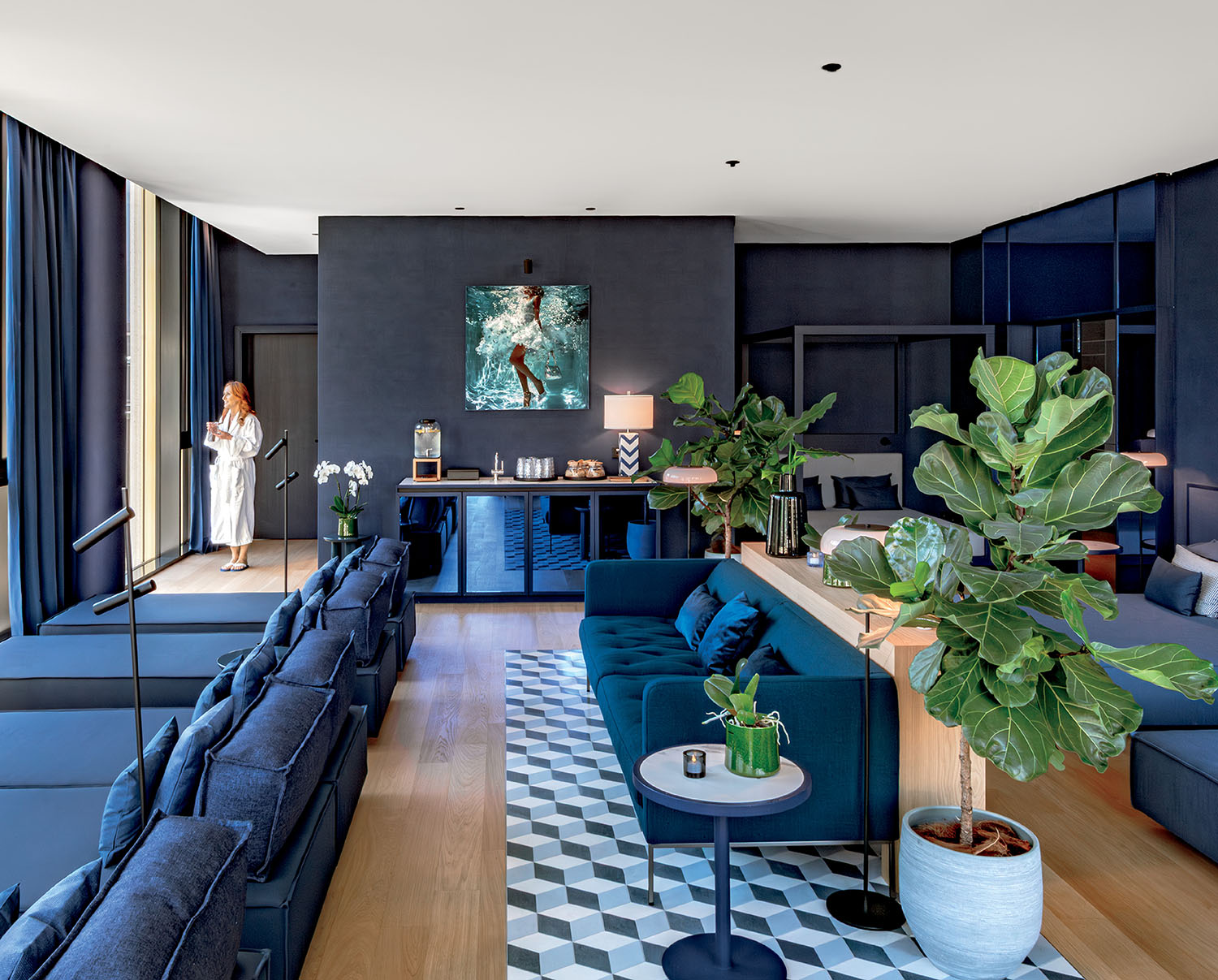 A modern living room with blue furniture, geometric rug, large potted plants, and a woman in a white robe standing by the window.