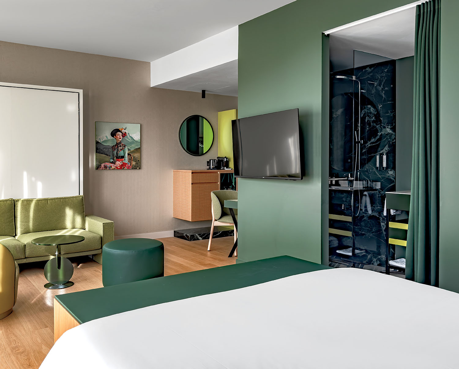 Modern hotel room with green and beige decor, a bed in the foreground, seating area, wall-mounted TV, kitchenette, and a glass-walled bathroom.
