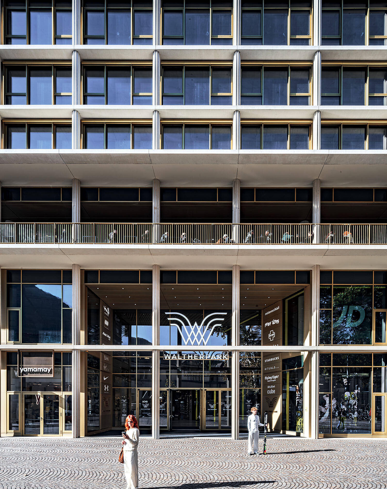 The entrance of a modern multi-story building labeled "WaltherPark," with glass doors, storefronts, and a few pedestrians in the foreground.