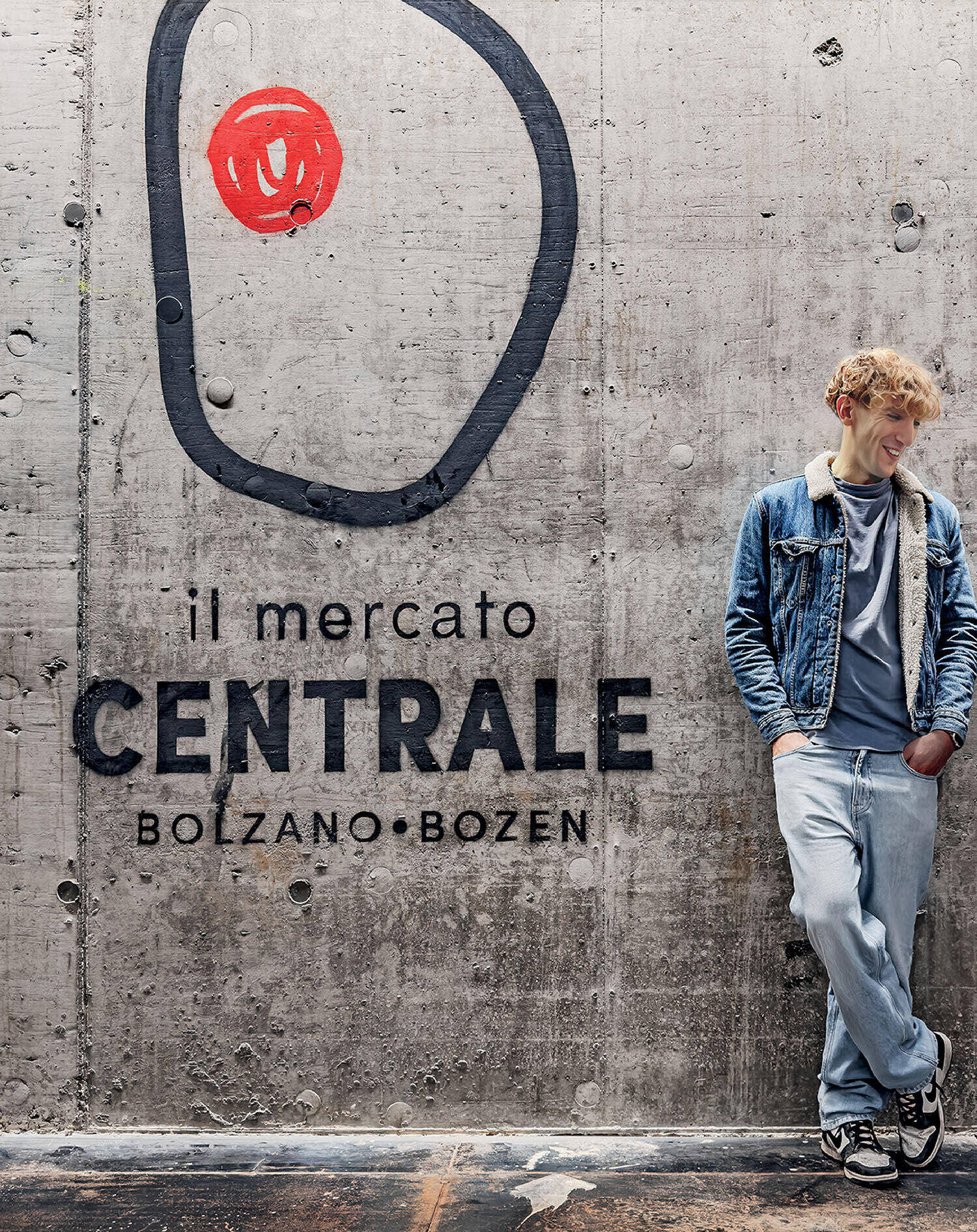 A young man in casual clothing leans against a concrete wall with "il mercato CENTRALE BOLZANO · BOZEN" and a red abstract design painted on it.