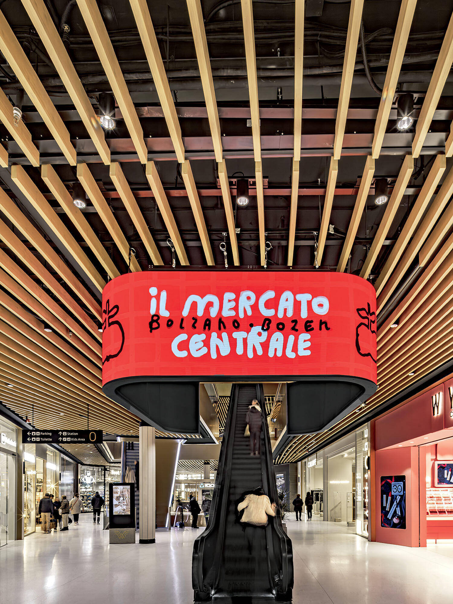 A large red sign reading "Il Mercato Centrale Bolzano Bozen" hangs above an escalator in a modern shopping mall with wooden ceiling slats and glass storefronts.