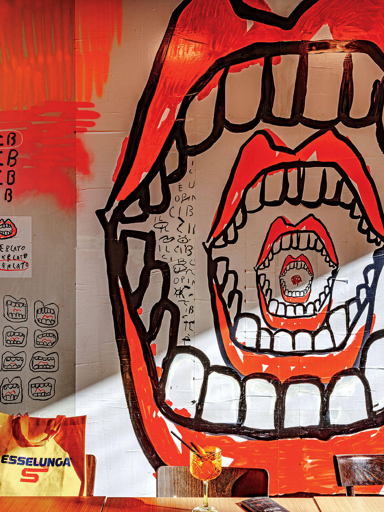 A mural of a large open mouth with smaller open mouths inside it, surrounded by abstract drawings and a shopping bag on the left; a drink is on the table in the foreground.