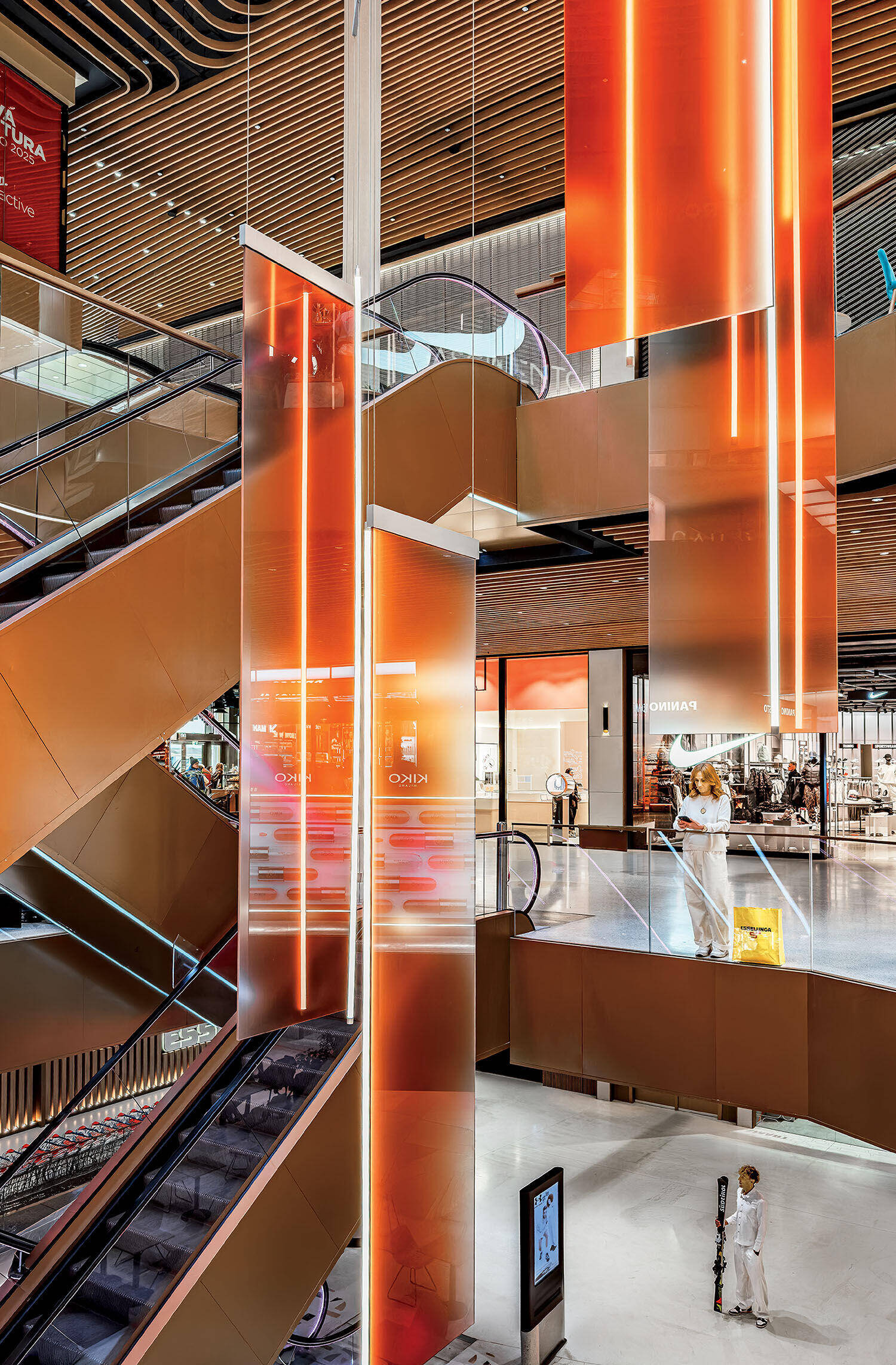 A modern shopping mall interior with orange illuminated panels, escalators, glass railings, and a few people walking and standing on different levels.