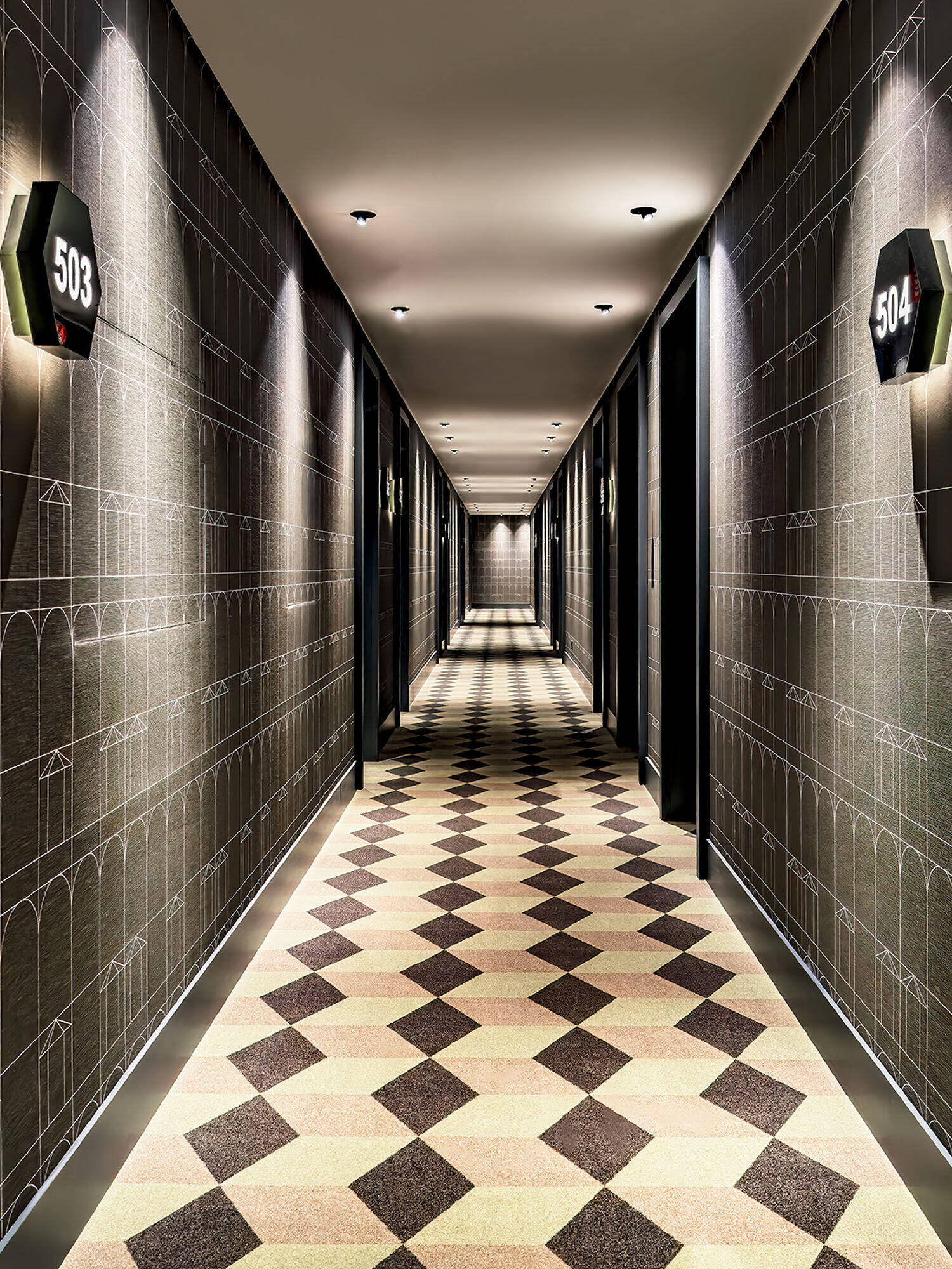 A well-lit hotel hallway with geometric patterned carpet, dark doors on both sides, and illuminated room number signs, including 503 and 504.