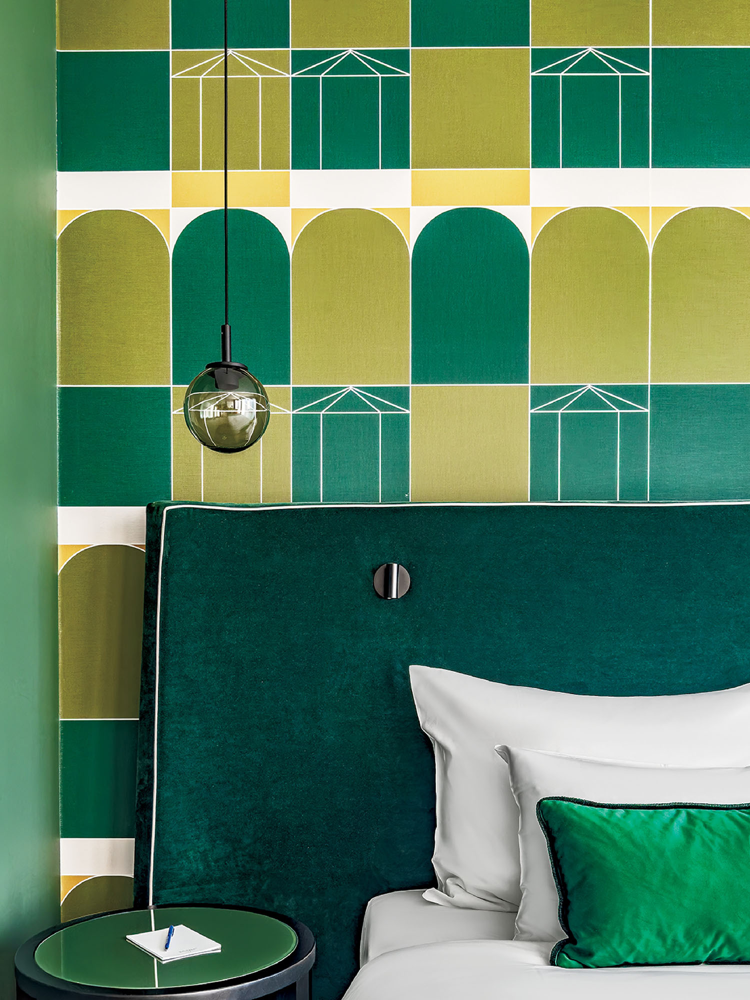 A modern bedroom with green geometric wallpaper, a velvet green headboard, white bedding with a green pillow, a small round side table, and a hanging round light fixture.