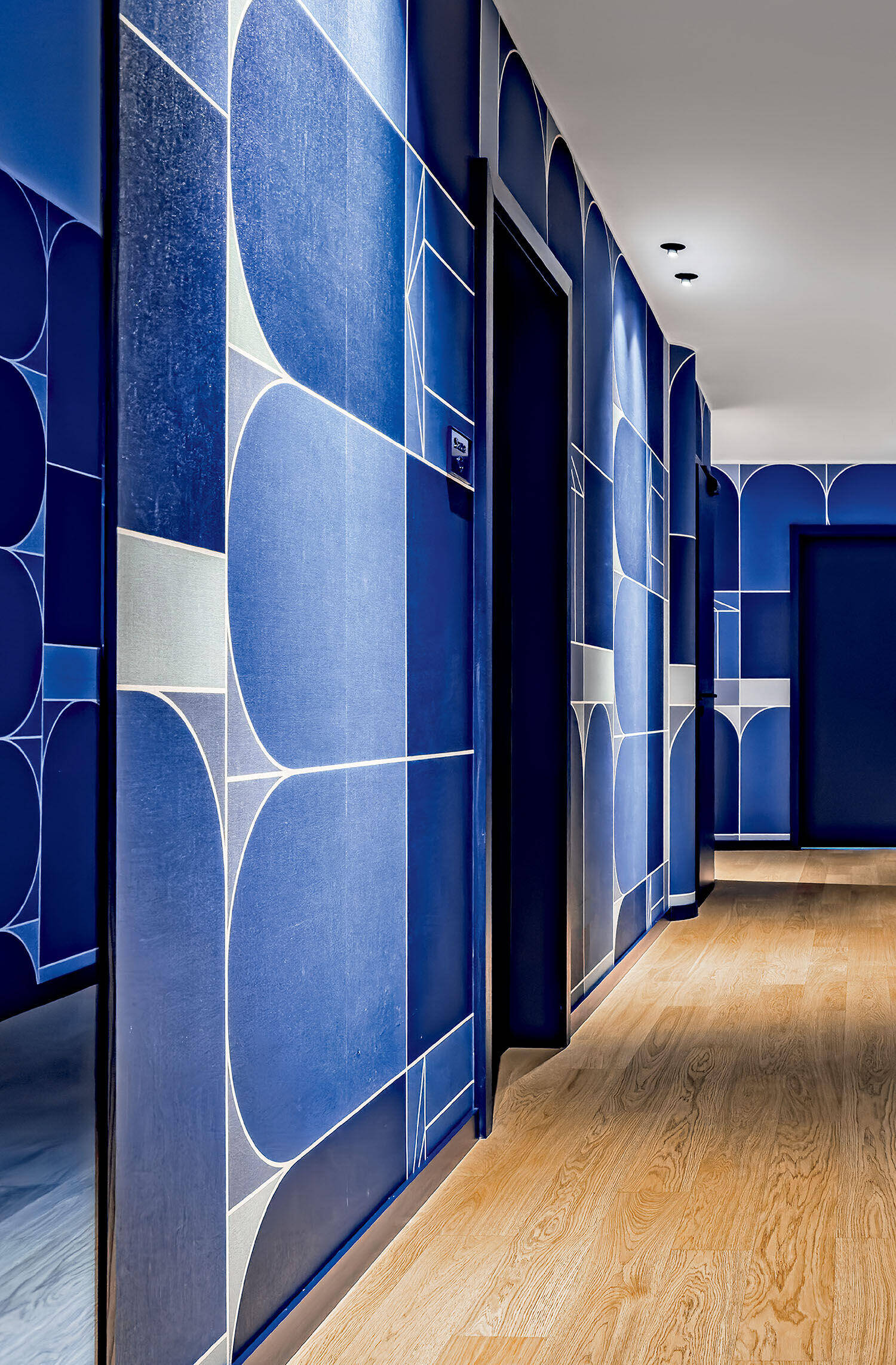 A hallway with blue geometric-patterned walls, light wood flooring, and several dark-framed doorways under recessed ceiling lights.