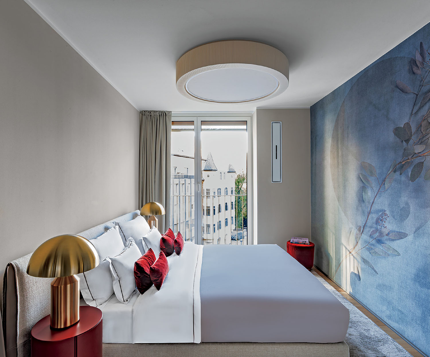 Modern bedroom with a large bed, gold lamps on red nightstands, blue accent wall with floral pattern, and a window overlooking city buildings.