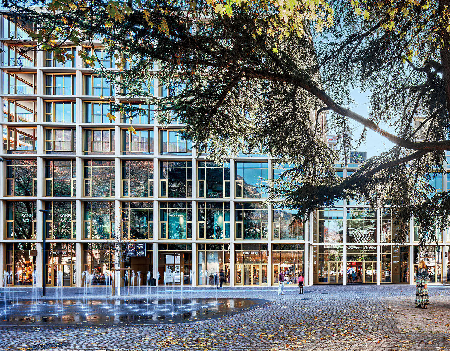 Modern glass and steel building with ground-floor shops, a cobblestone plaza, water fountains, and trees in the foreground under a clear sky.