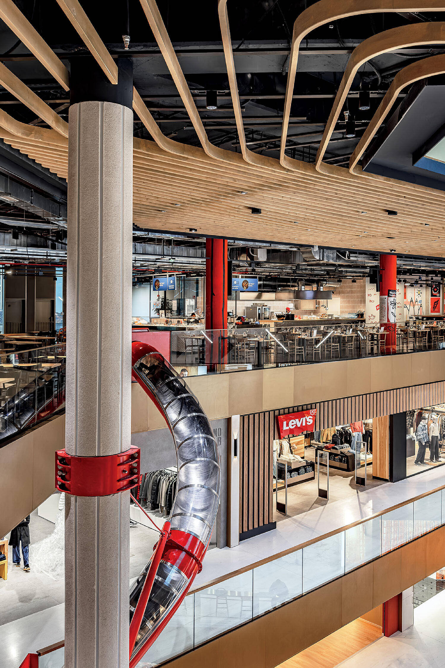 Interior of a modern shopping mall with wooden ceiling panels, a metal slide winding around a column, glass railings, and a Levi's store on the lower level.
