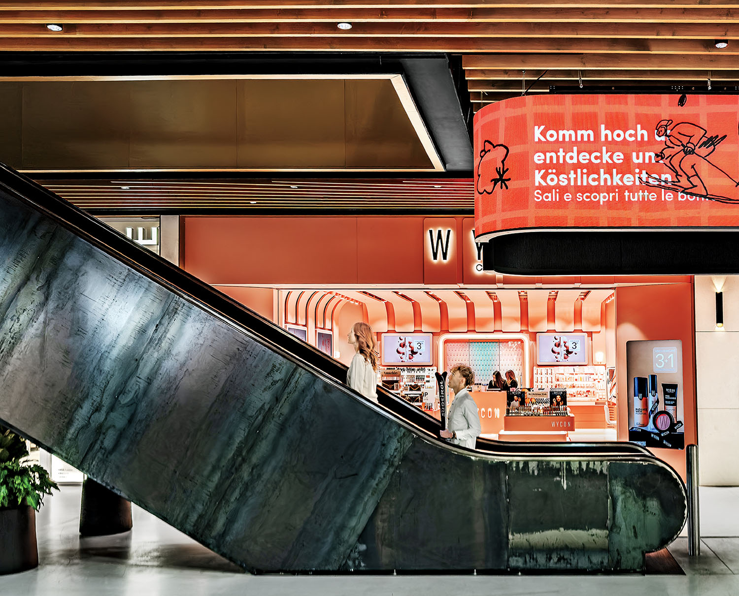 Two people ride an escalator past a modern orange shop with German and Italian signage inside a contemporary building.