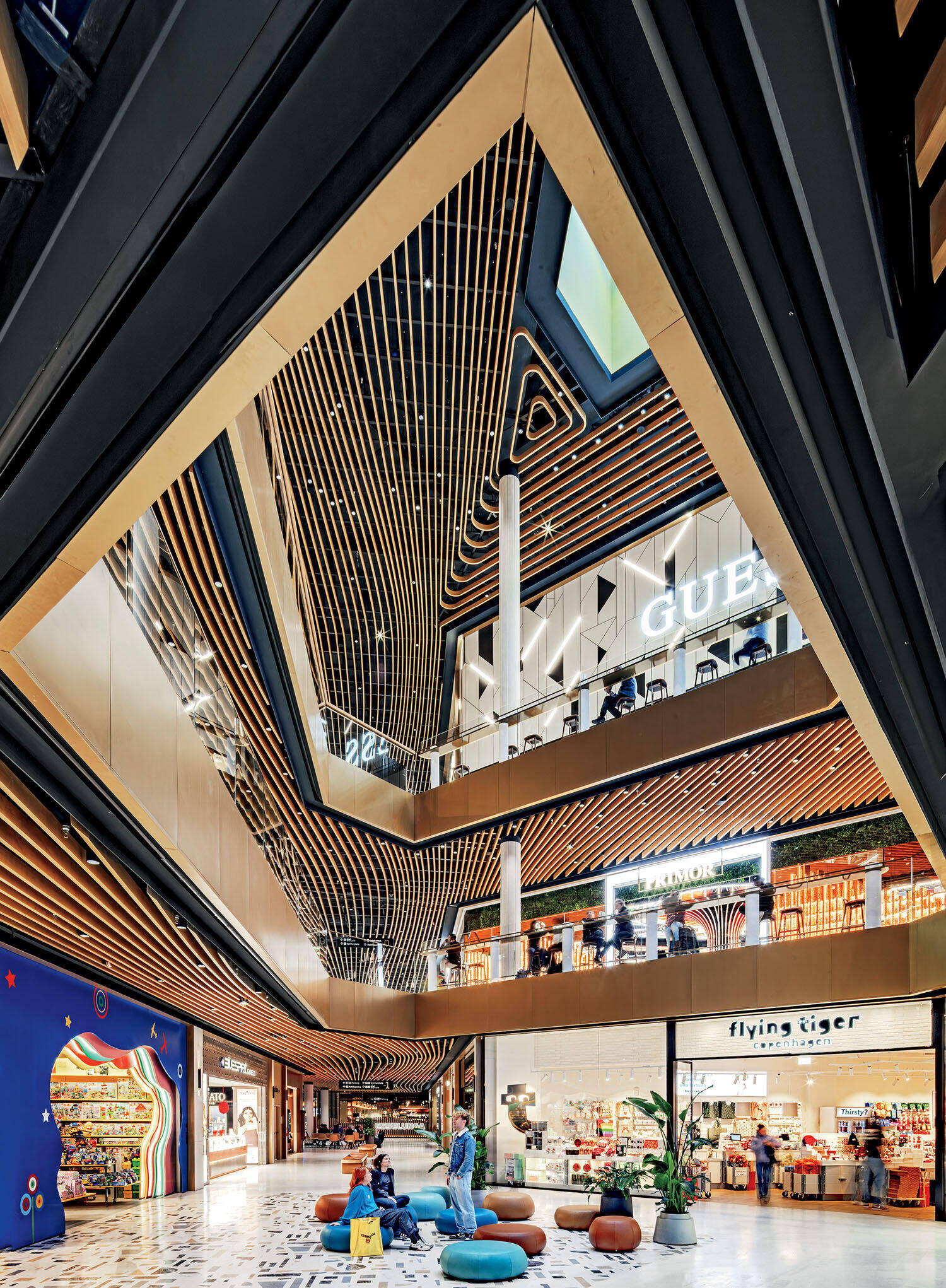 A spacious modern shopping mall with multiple floors, geometric wooden ceiling accents, and stores including Guess and Flying Tiger; people are seated on colorful ottomans.
