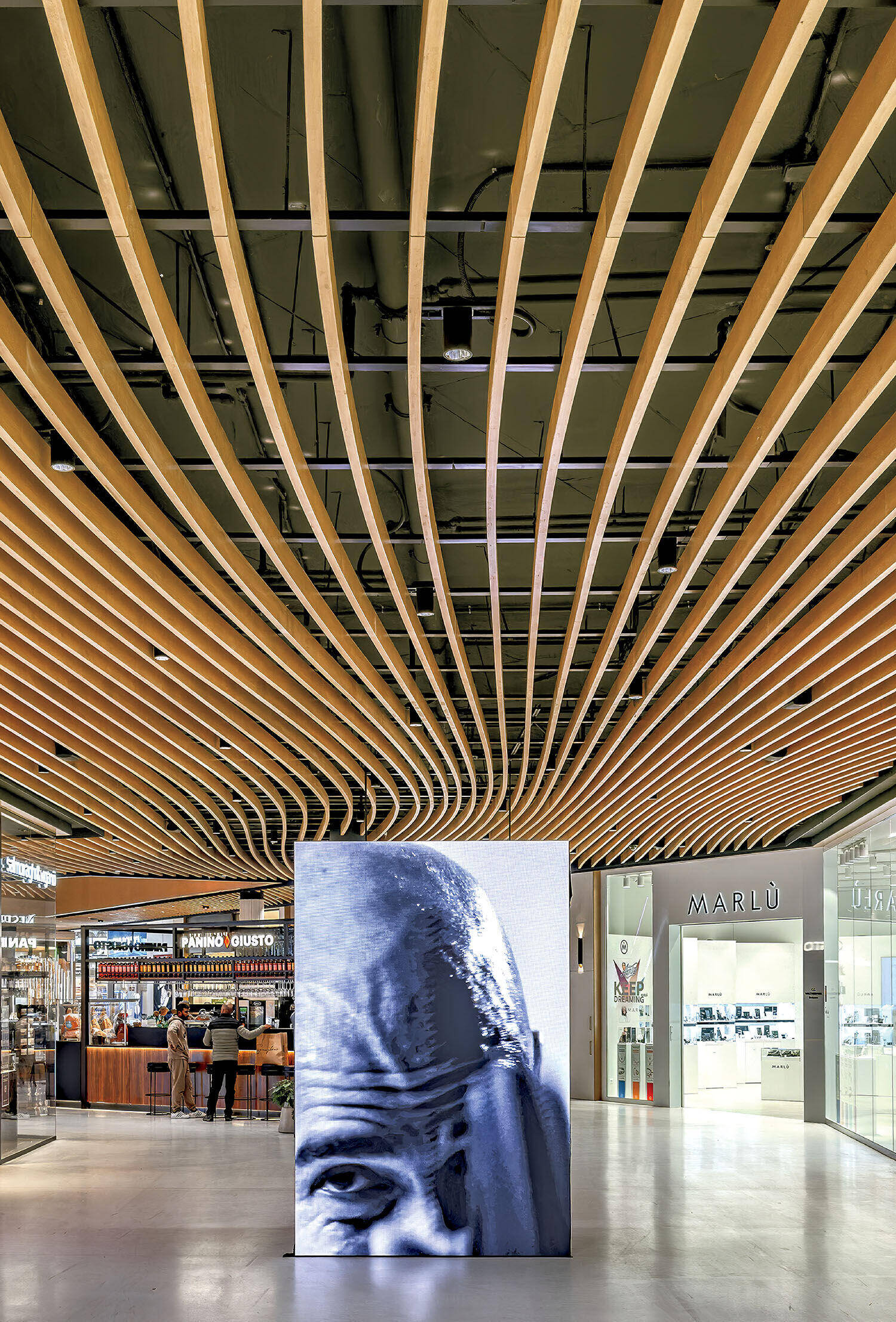 Large digital screen displaying a close-up image of a face in a modern shopping mall with curved wooden ceiling slats and various store fronts.