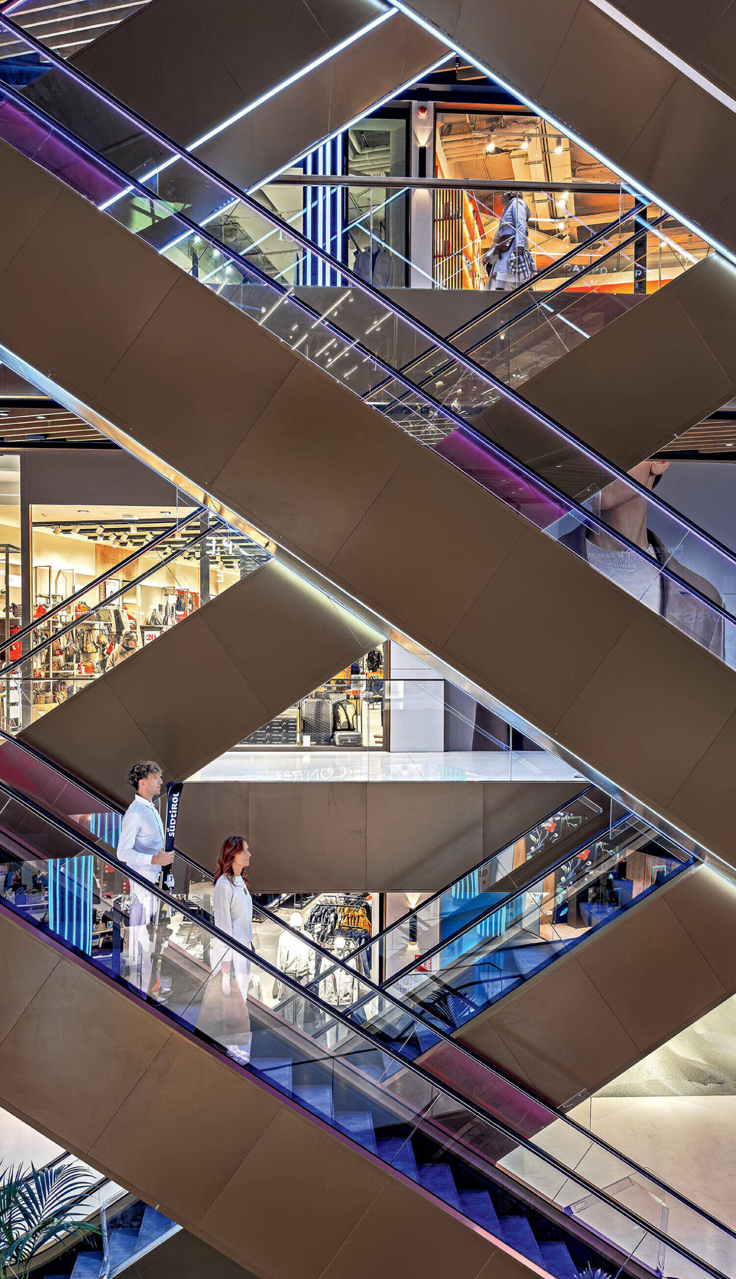 Several people ride escalators in opposite directions inside a modern, well-lit building with geometric architecture and visible retail spaces.