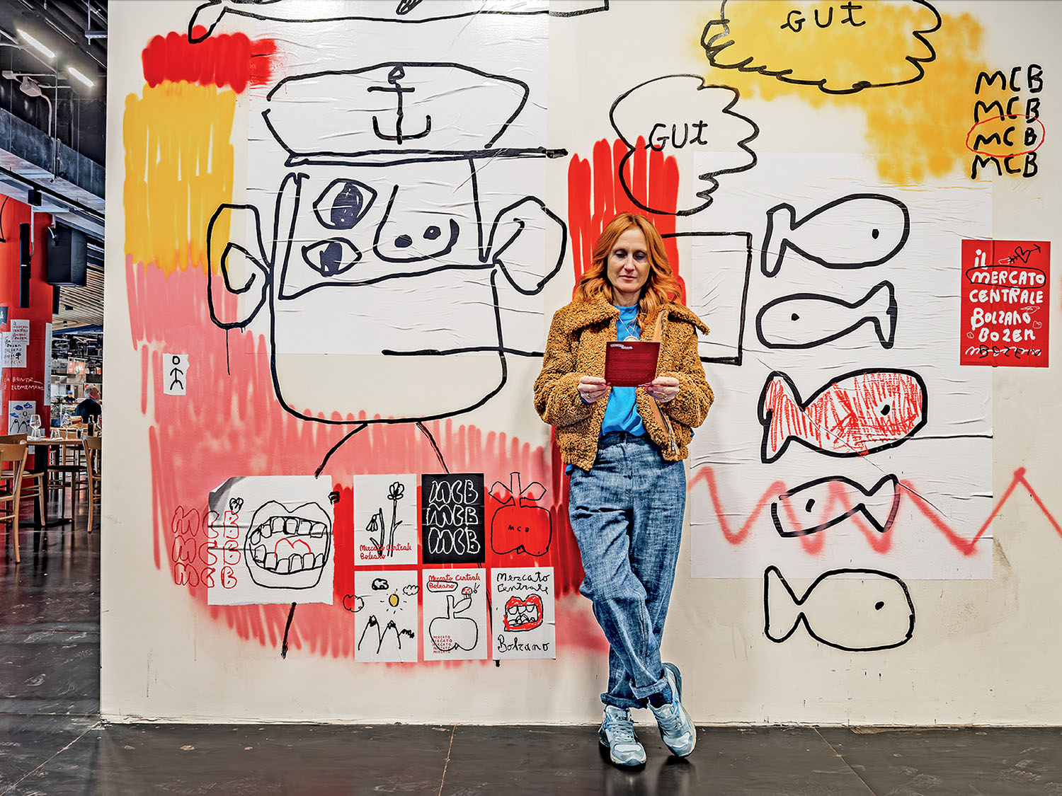 A woman in casual clothes holds a red mug while standing against a wall covered with colorful, childlike drawings and sketches in an indoor public space.