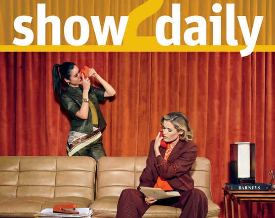 Two women in a retro-style room; one stands on a sofa speaking on a red phone, the other sits on the sofa holding a phone. A stack of magazines and curtains are in the background.