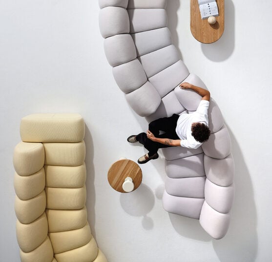 A person sits on a curved, light gray modular sofa beside a coffee table and a wall-mounted shelf, viewed from above. Text at the top reads "NeoCon show daily 3 INTERIOR DESIGN.