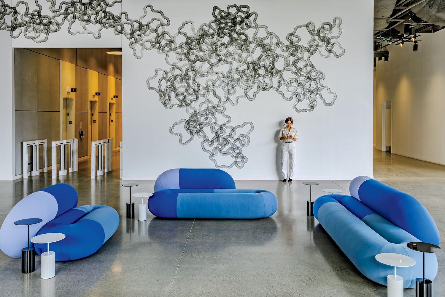 Modern lobby with three blue sofas, small white tables, abstract wall art, and a person standing by the wall using a phone. Elevator doors and turnstiles are visible in the background.
