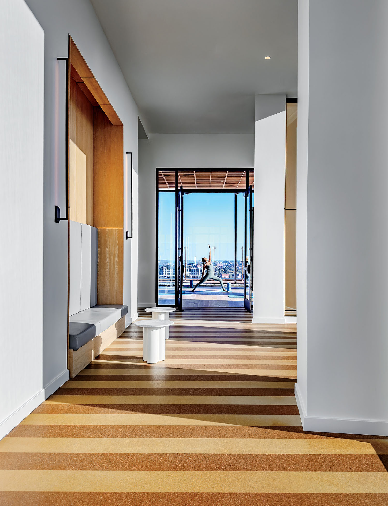 A modern hallway with striped flooring, a built-in bench, and glass doors opens to a balcony where a person is jumping with arms raised in the distance.