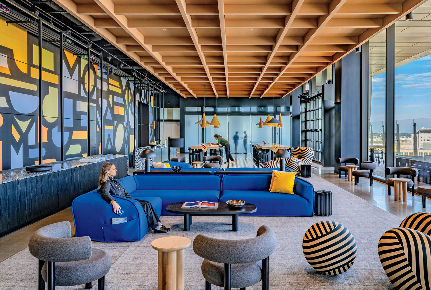 Modern lounge with blue and gray seating, geometric wall art, round striped stools, large windows, and a person sitting on a sofa.