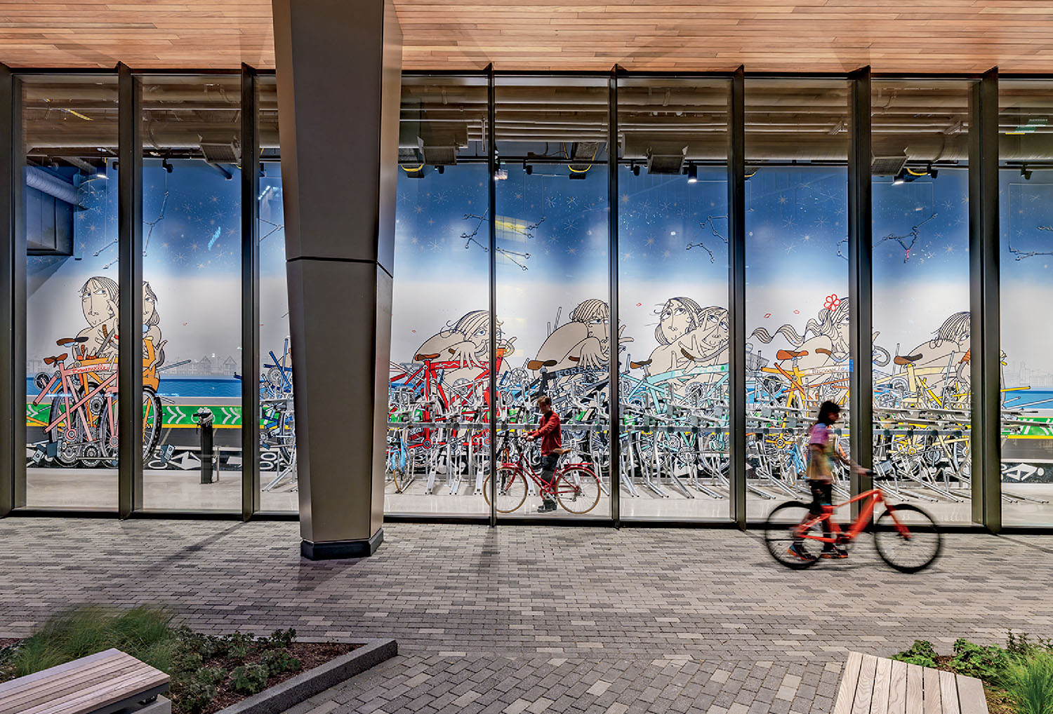 Two people ride bikes in front of a building with large glass windows displaying a colorful mural of children riding bicycles.