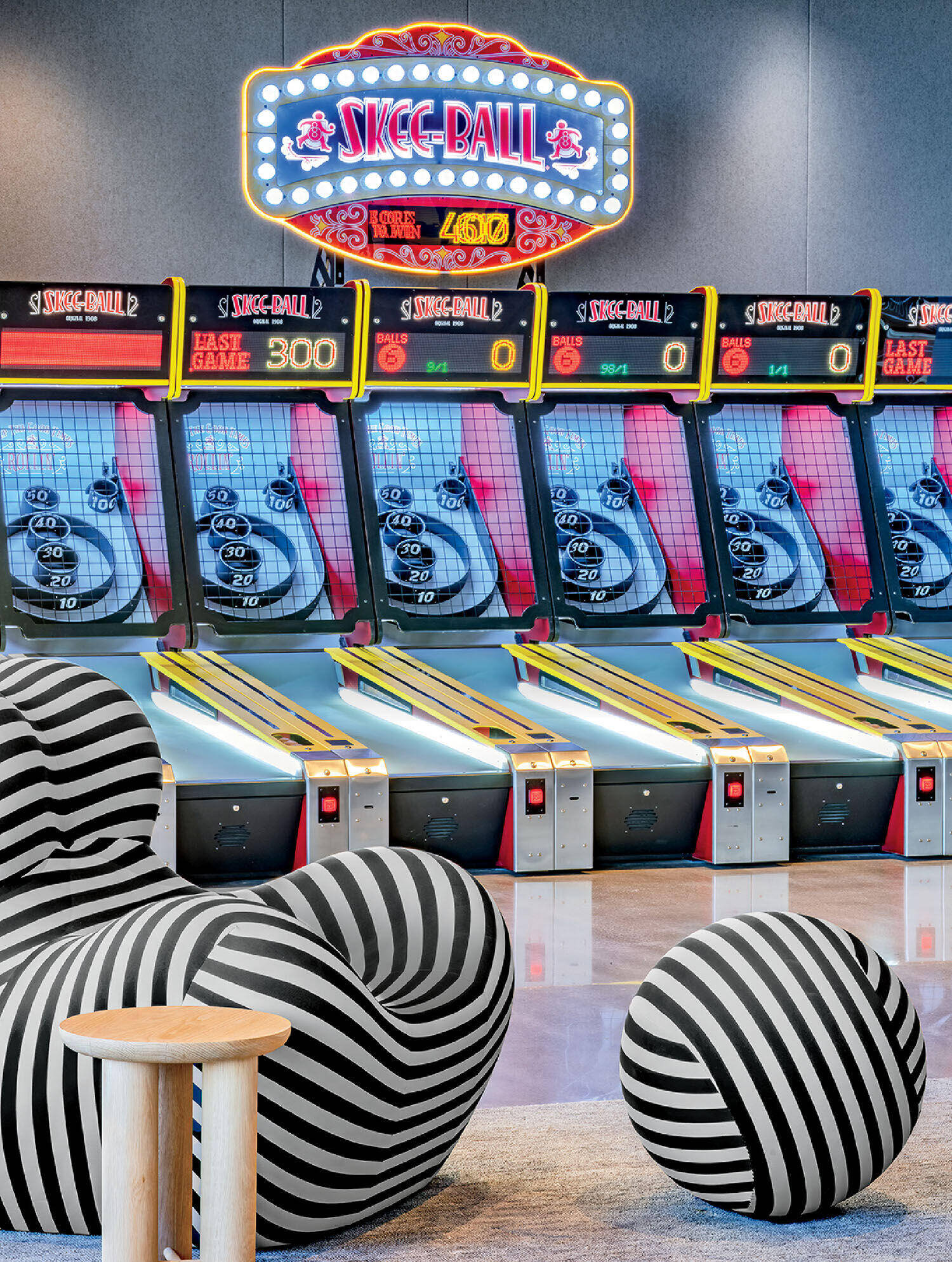 A row of Skee-Ball arcade machines is lined up against a wall, with striped modern seating and a small wooden stool in the foreground.