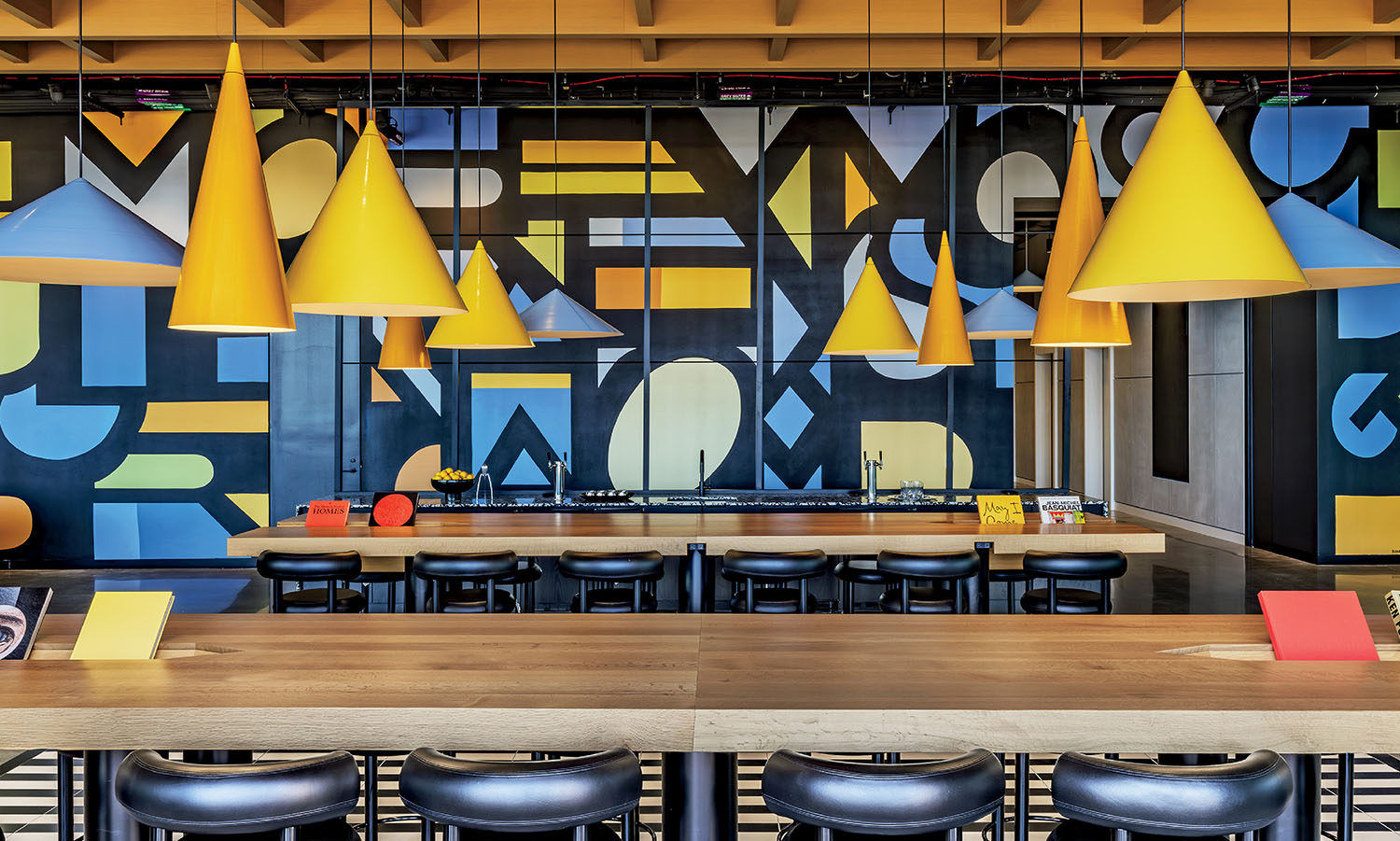 A modern communal workspace with long wooden tables, black stools, yellow cone-shaped pendant lights, and a colorful geometric mural on the back wall.