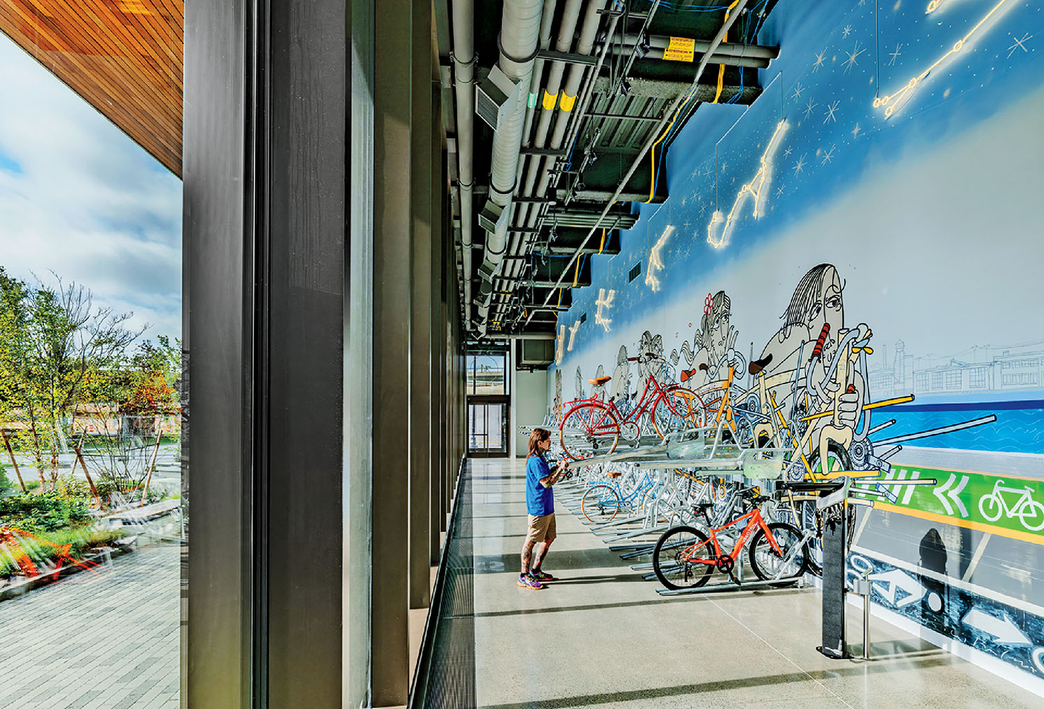 A person stands in a modern hallway with floor-to-ceiling windows, observing a colorful mural of bicycles and stars; several bikes are parked along the wall.