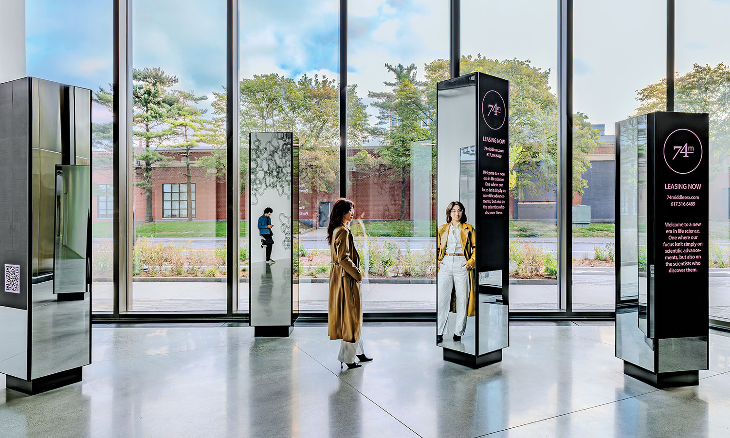 A woman stands in a modern, glass-walled gallery space, looking at interactive digital displays with text and images. Large windows reveal trees and a brick building outside.