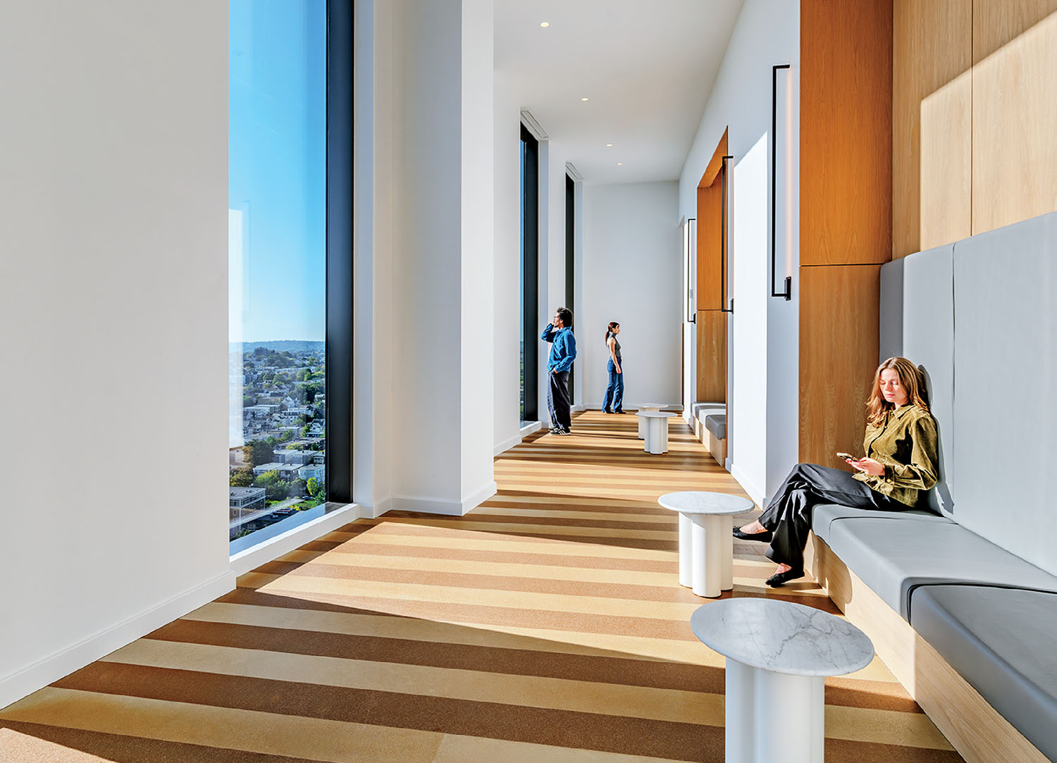 A modern, sunlit hallway with floor-to-ceiling windows, striped flooring, and built-in seating; three people are present, one seated and two standing.