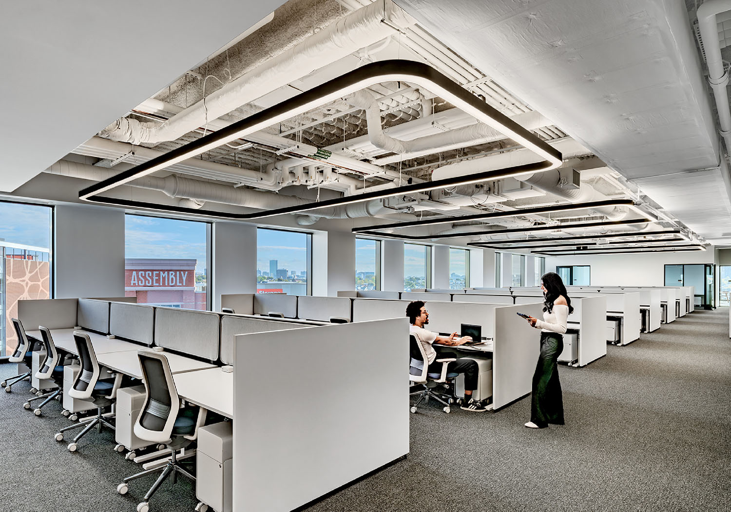 Modern open-plan office with rows of desks and chairs, two people conversing near workstations, large windows providing natural light.