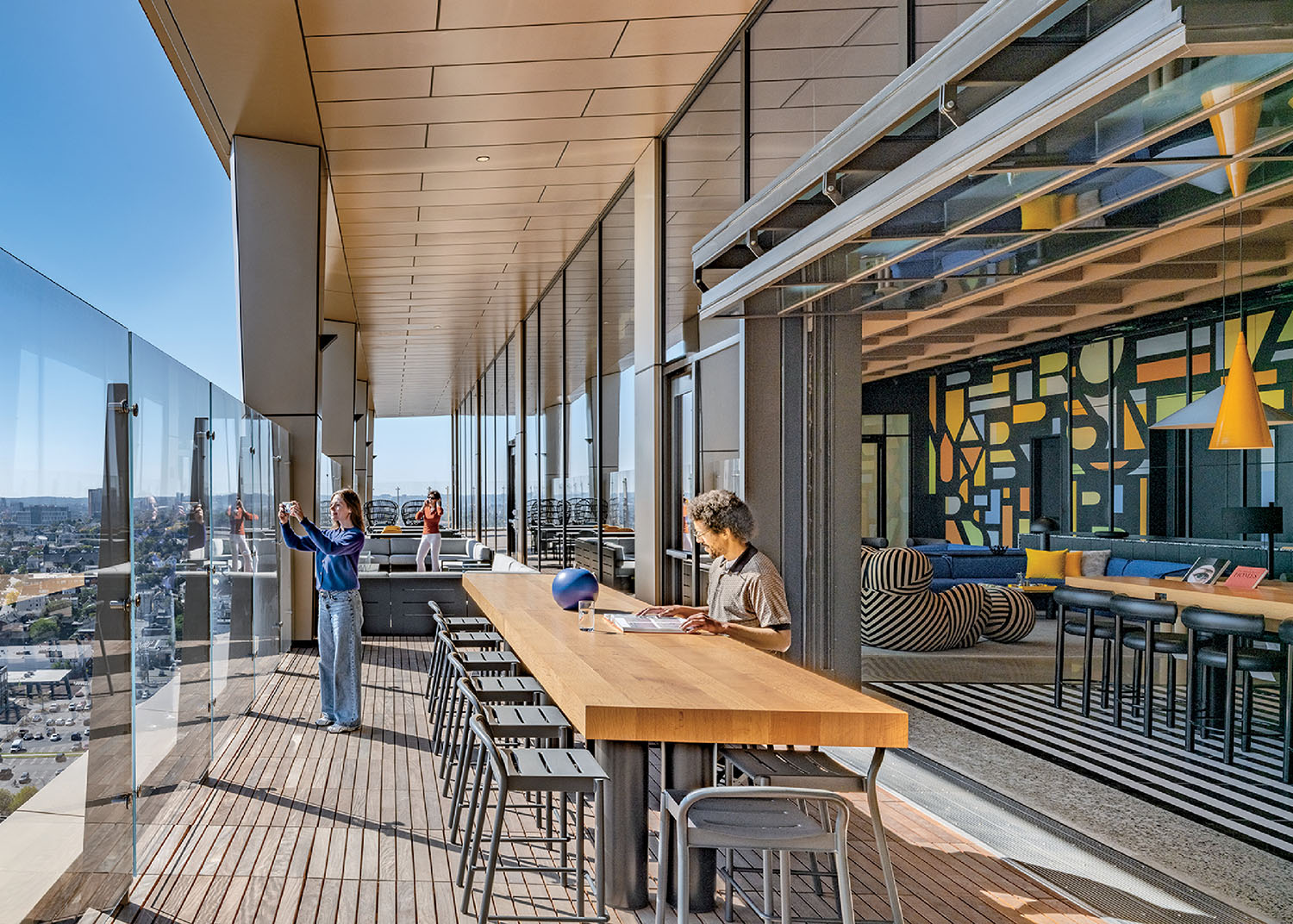A modern rooftop terrace with glass railing features people sitting at tables and one person taking a photo, adjacent to a colorful indoor lounge area.