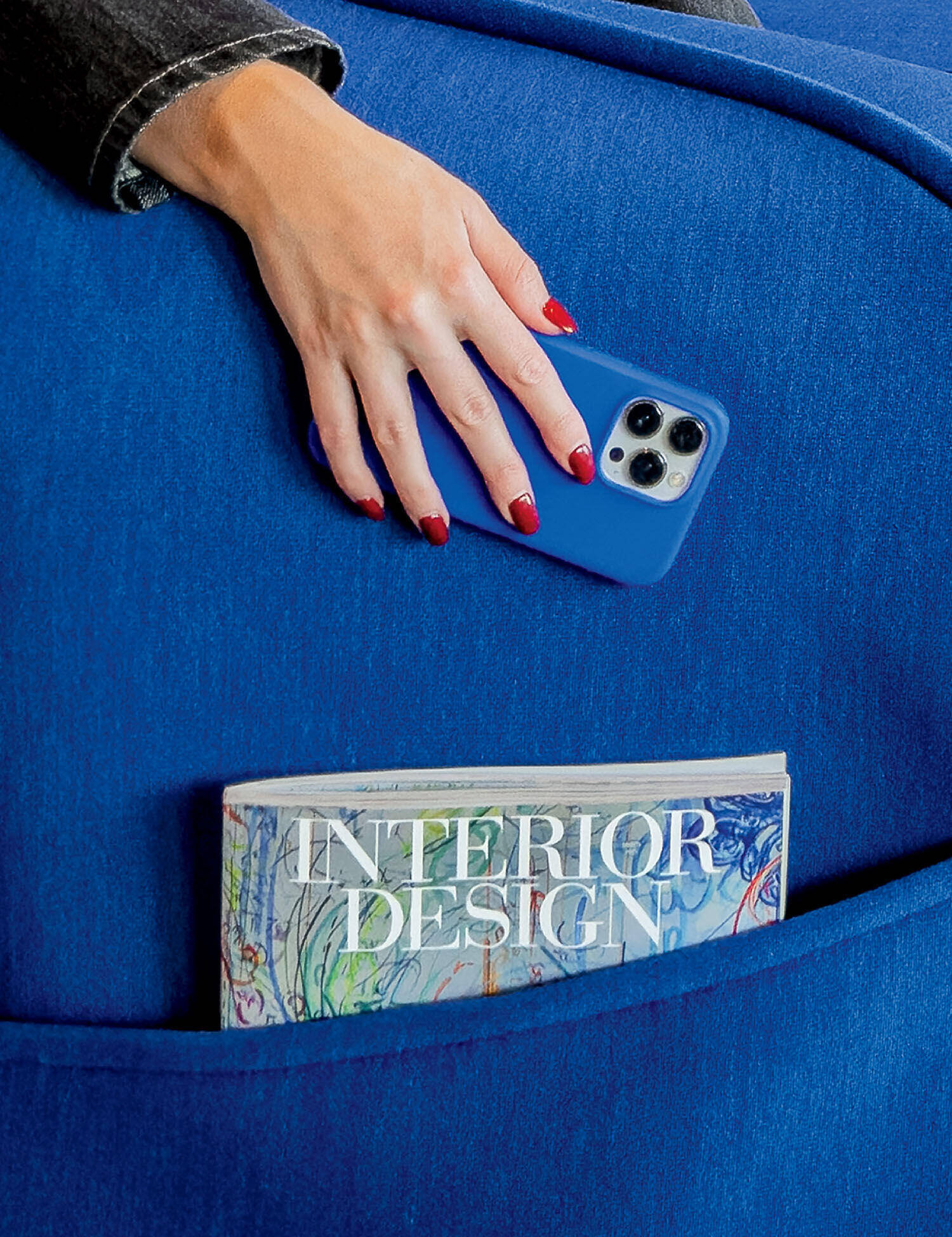 A hand with red nail polish holds a blue smartphone on a blue couch, with a magazine titled "Interior Design" in the couch pocket.