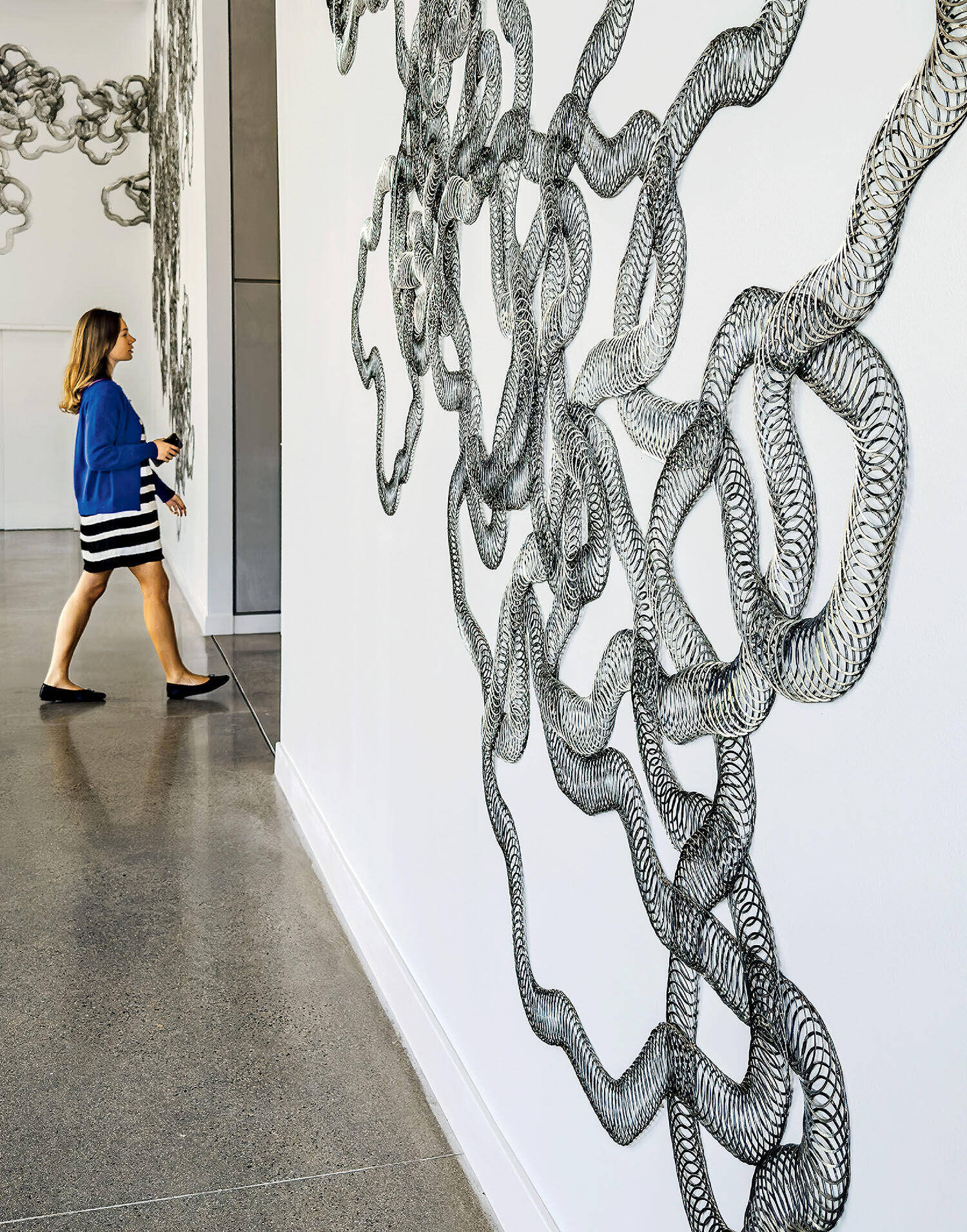 A woman walks through a gallery with large, intricate black and white abstract wall art featuring intertwined, looping lines.