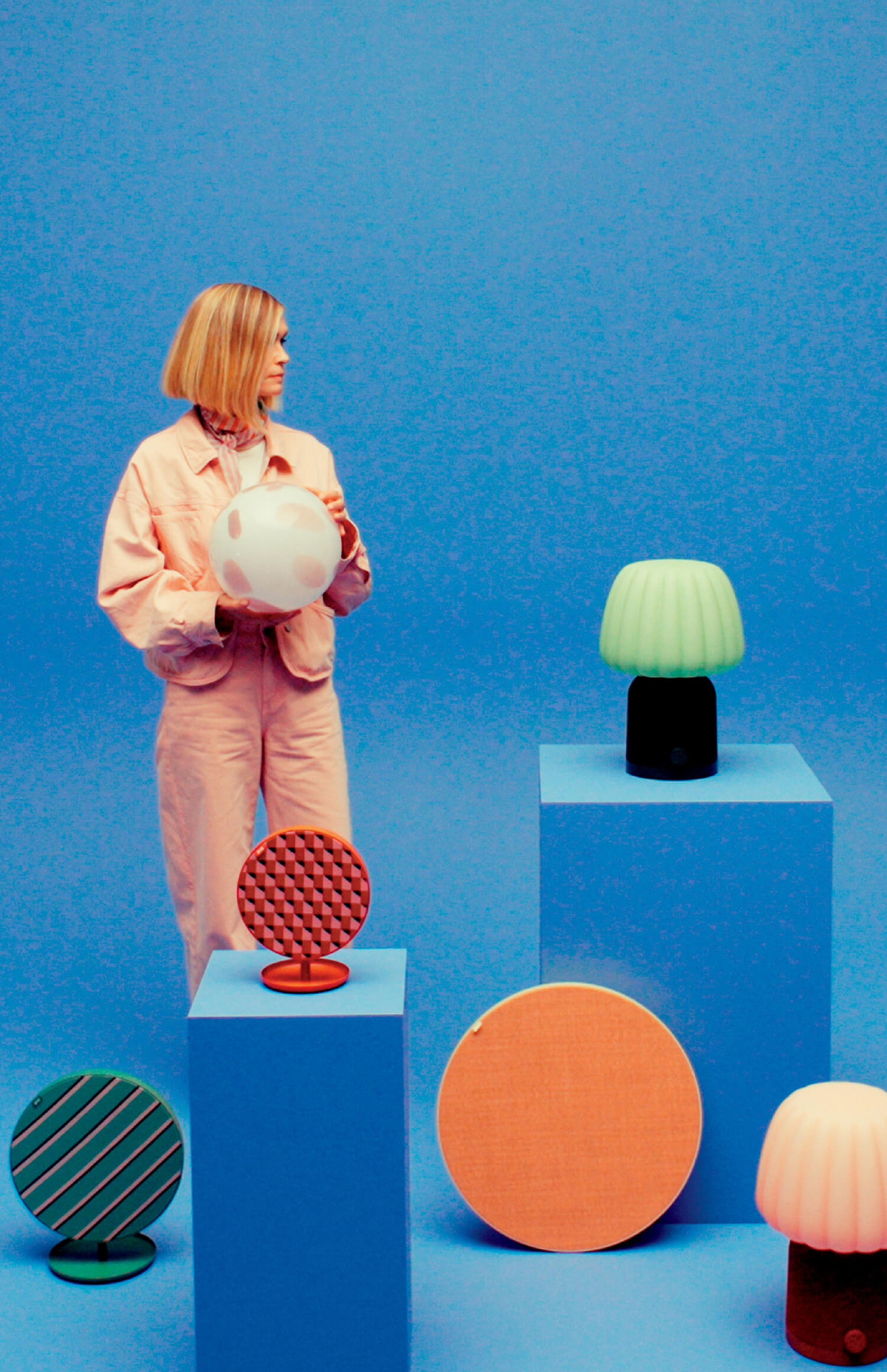 A woman in a light pink jumpsuit stands among blue display pedestals holding a round white lamp, with various colorful lamps and objects arranged around her, all against a blue background.