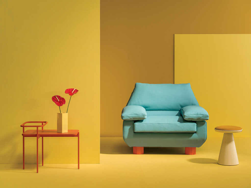 A blue armchair with wooden legs sits between a small orange table holding red flowers and a round white and yellow stool, set against a yellow background.